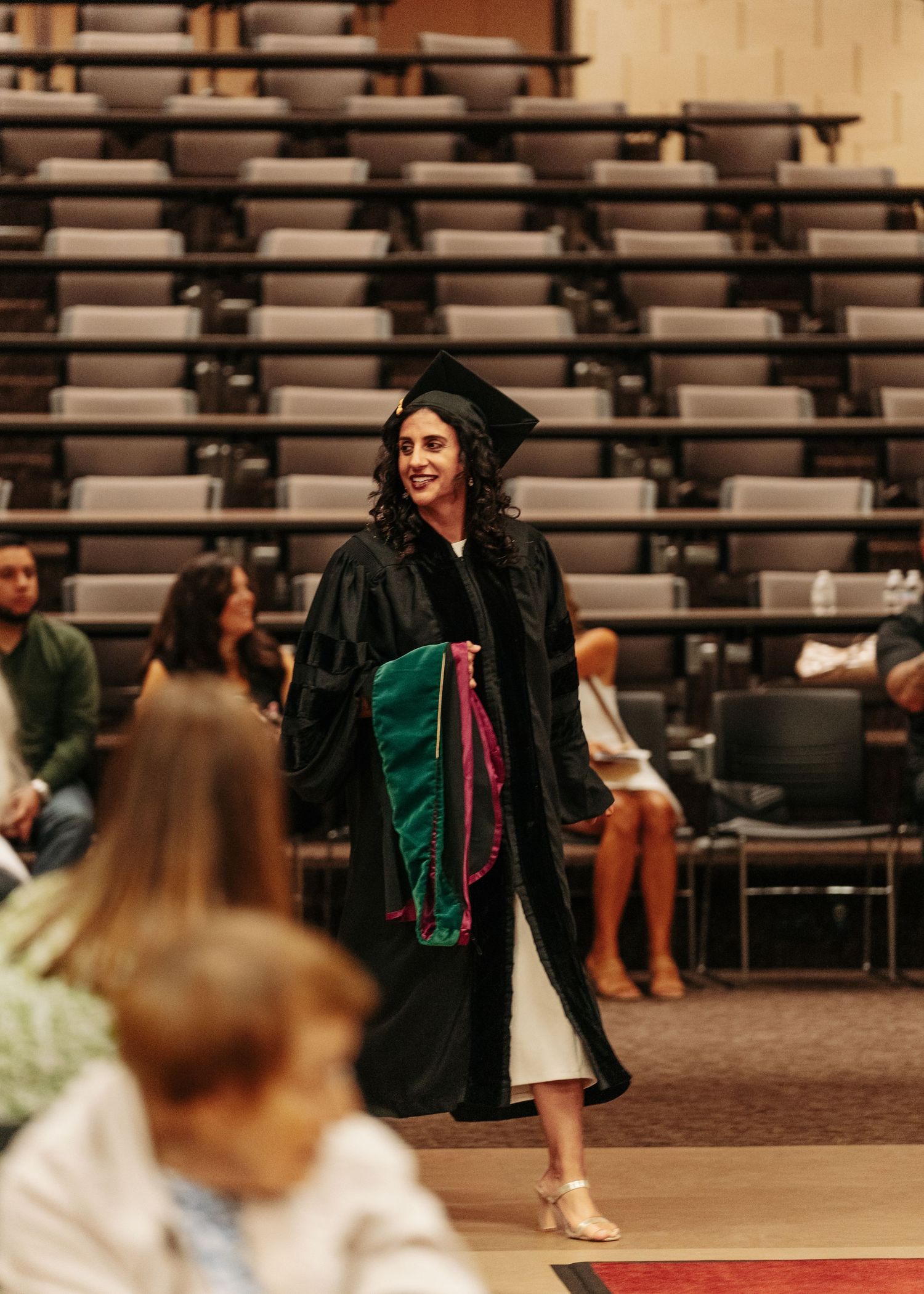 Graduate in black cap and gown smiling while walking during commencement ceremony in an auditorium.
