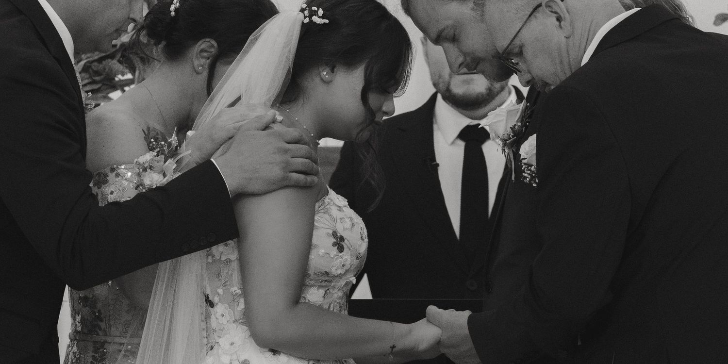 Black and white photo of couple holding hands in prayer with officiant at wedding altar.