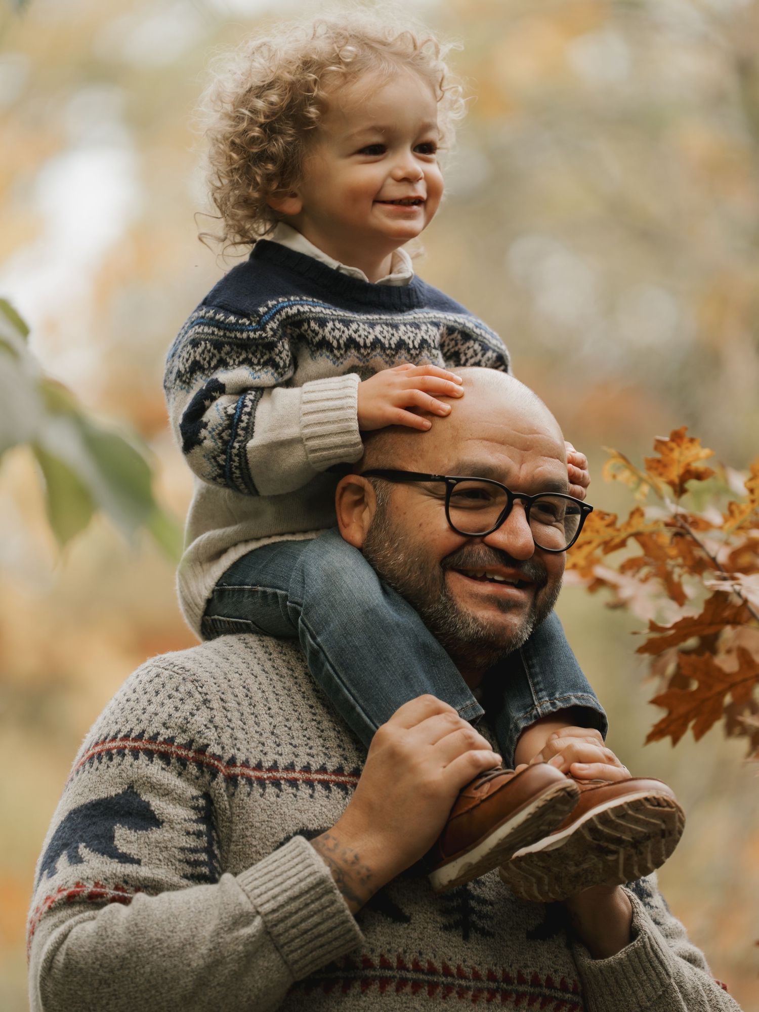 Smiling father with toddler sitting on shoulders among fall foliage.