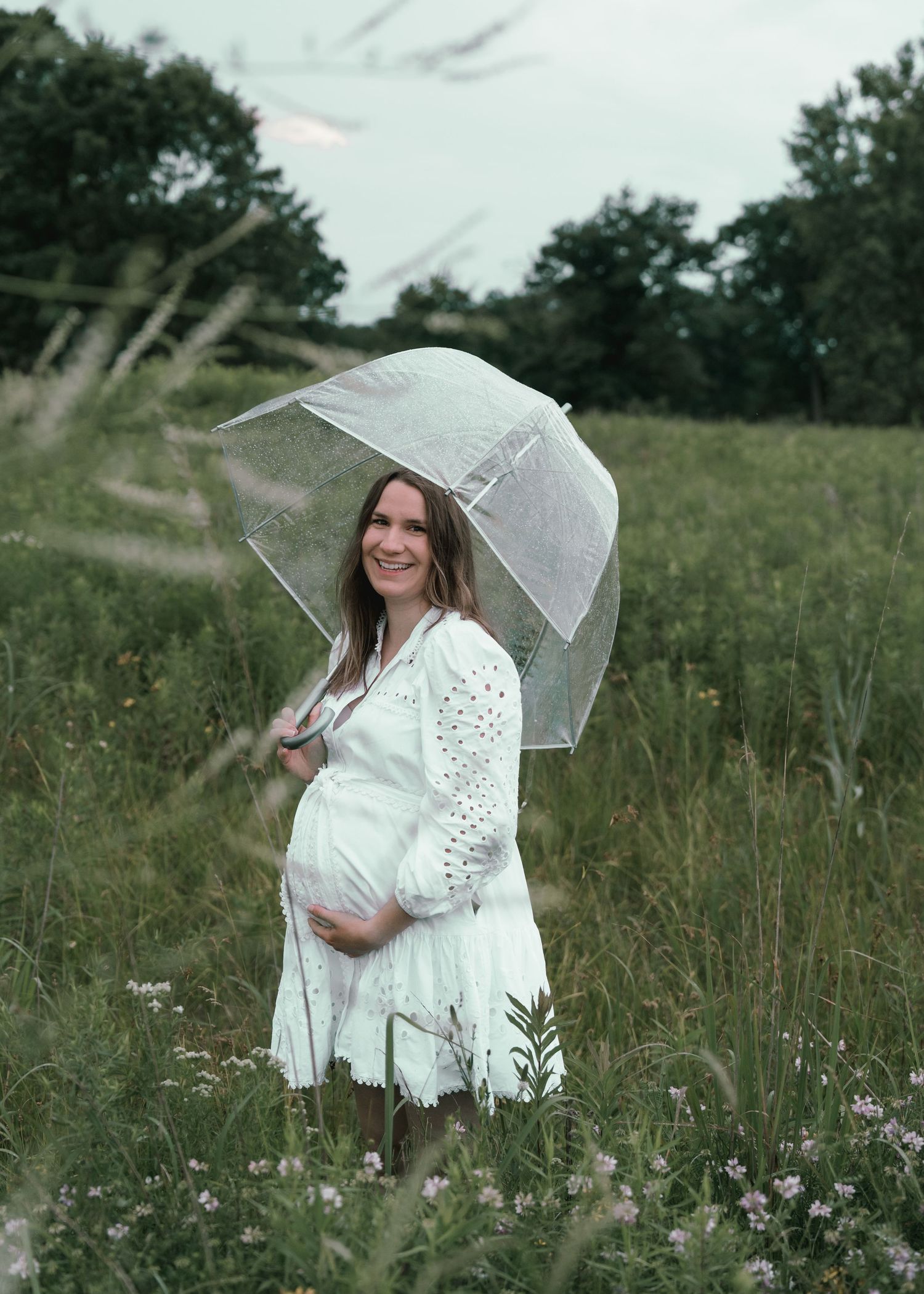 Smiling pregnant mother in white dress holding a clear umbrella in a lush green meadow.