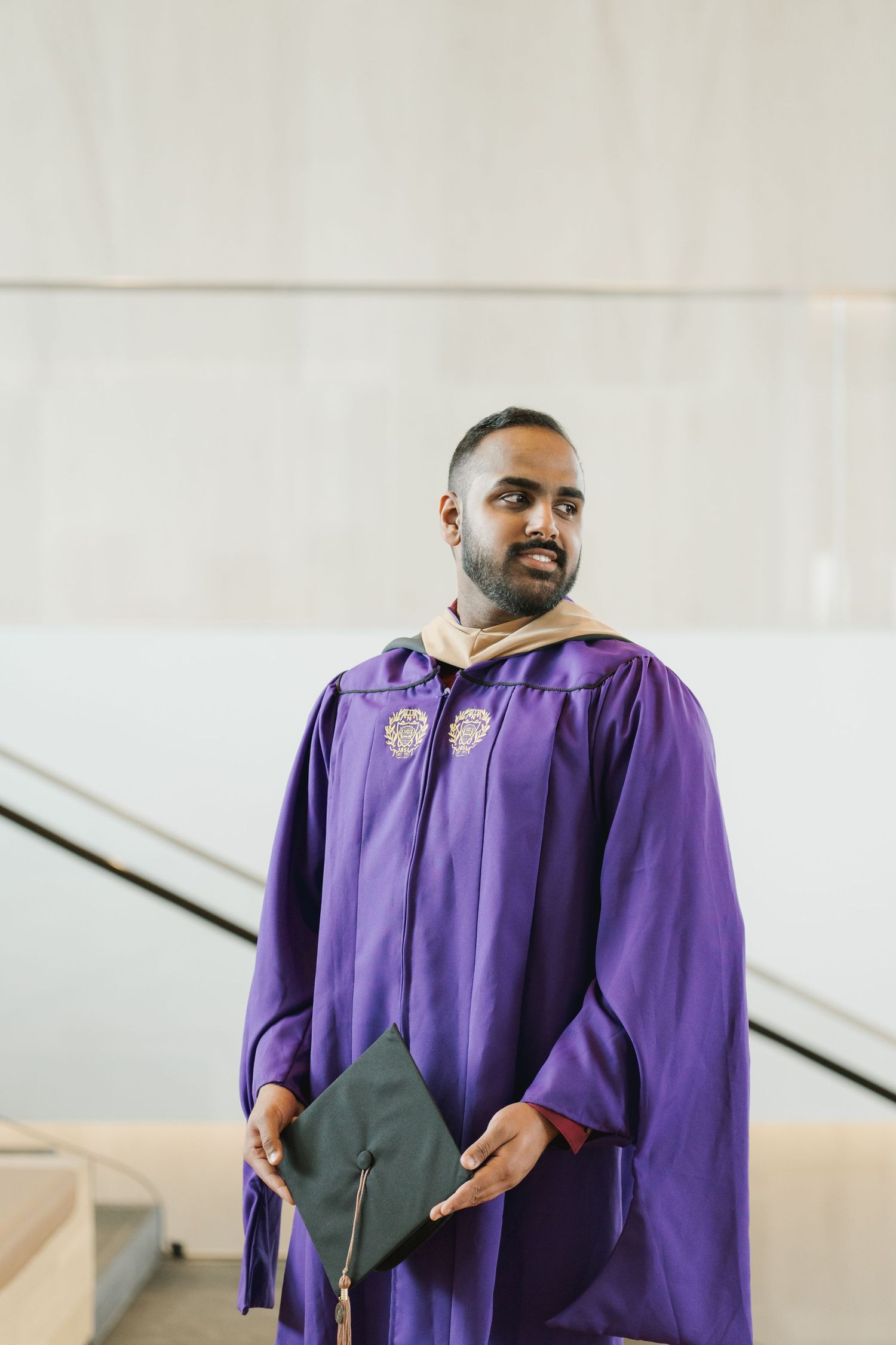 Man in purple graduation gown holding a cap in a modern building interior.