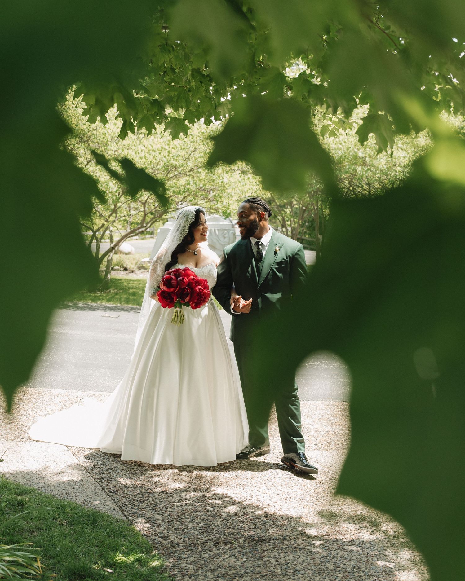 Bride in white gown with red bouquet and groom in green suit walk a garden path, framed by lush green leaves.