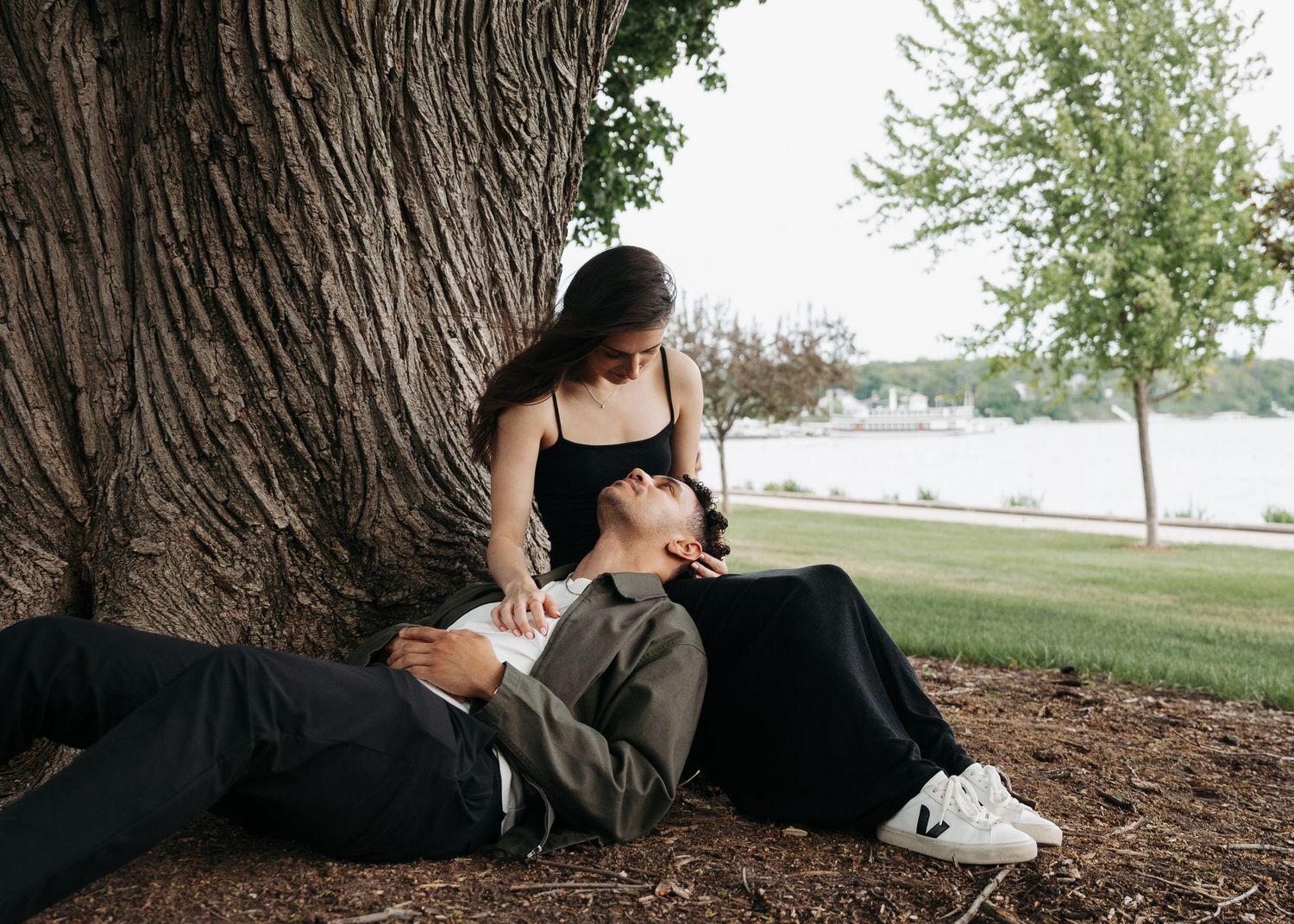 Couple sharing a romantic moment under a large tree in a park near a lake, woman in black top, man lying down.