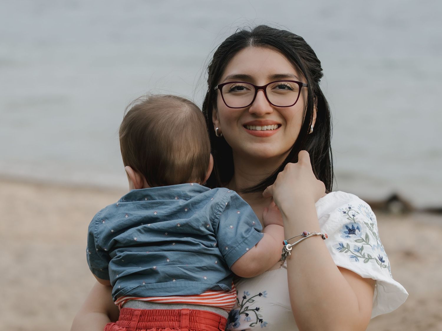 Young woman with glasses smiling at beach while holding a baby in a denim outfit on a sunny day.