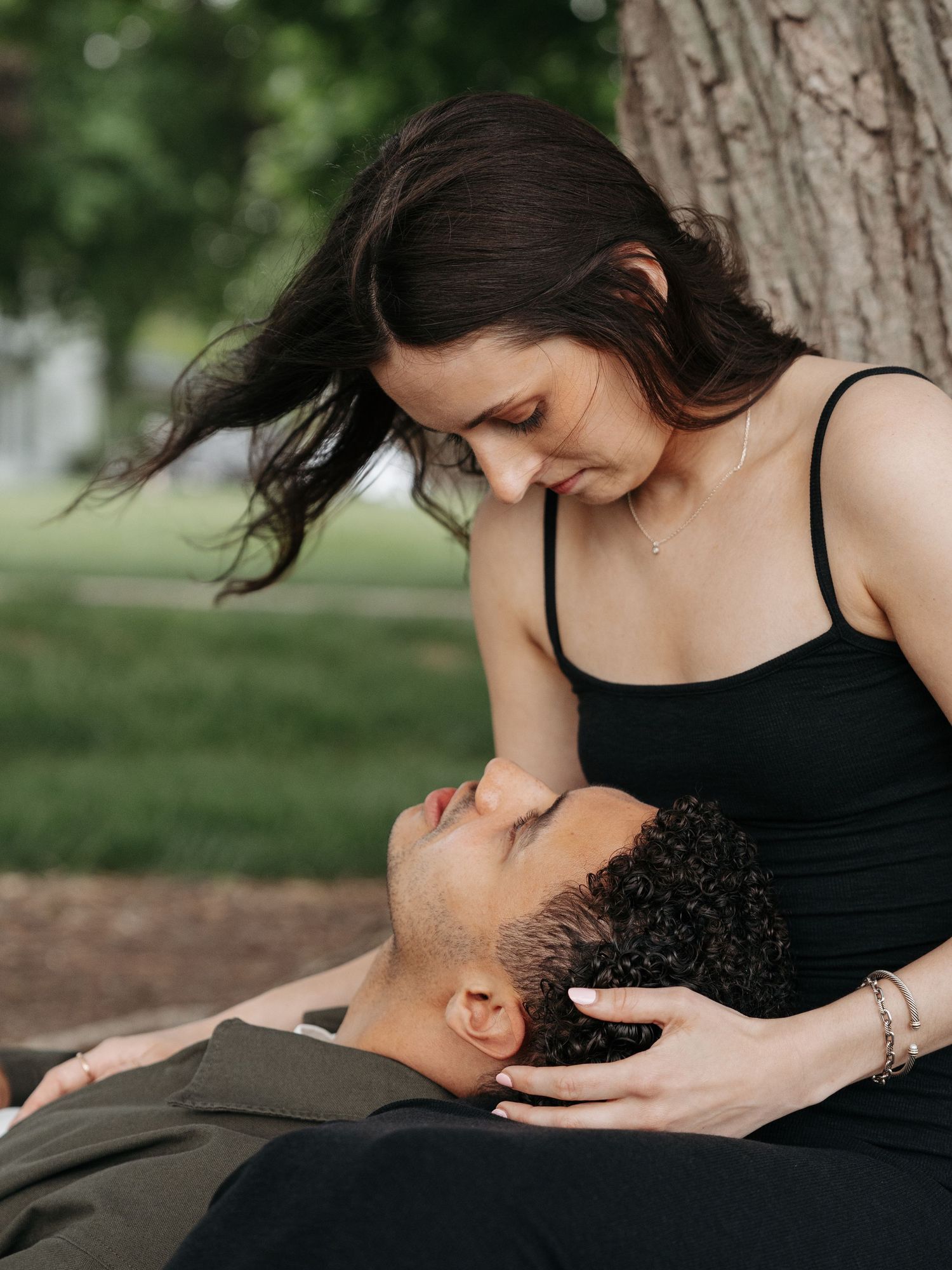 A couple shares an intimate moment outdoors, woman in black dress cradling man's head in her lap near a tree.