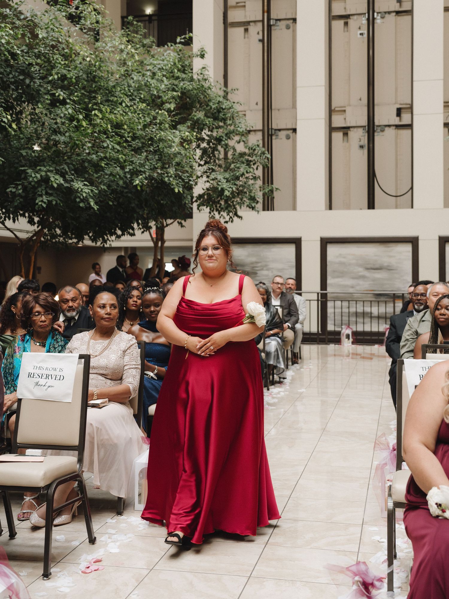 Bridesmaid in red satin gown walks down wedding ceremony aisle in elegant indoor venue.