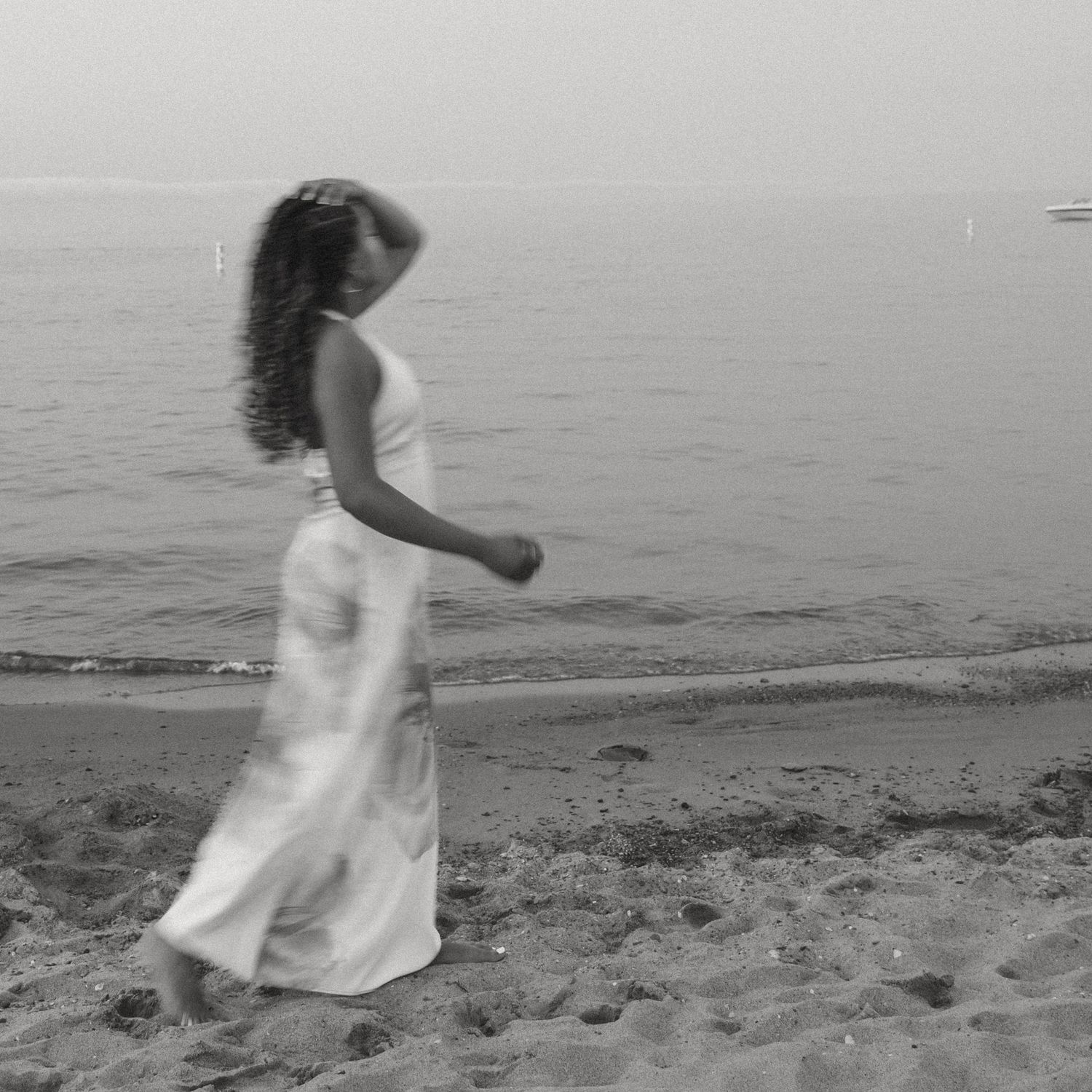 Black and white photo of a woman in a flowing white dress walking barefoot along a sandy beach near calm water.