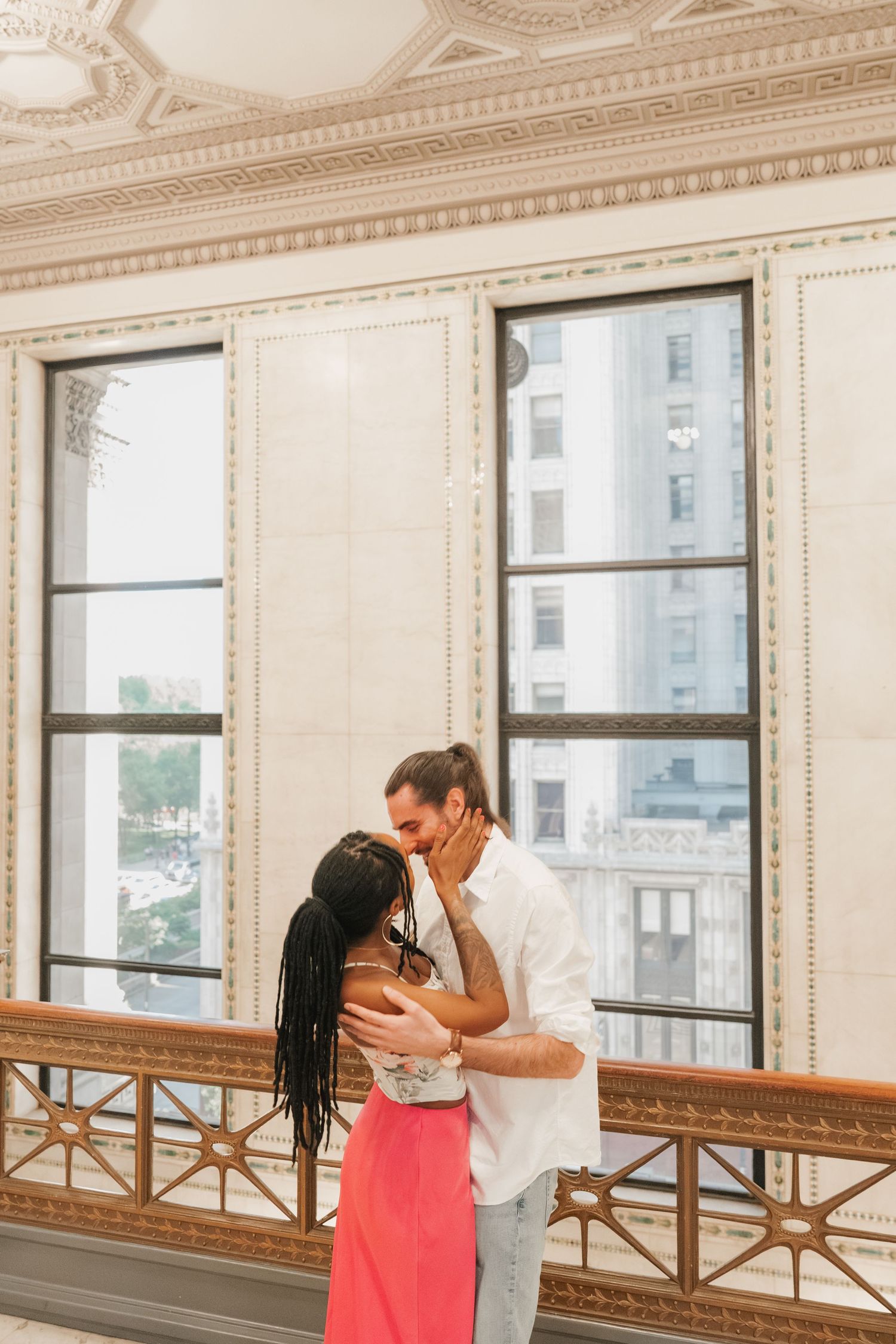 Couple embracing and kissing near large windows inside a grand historic building.