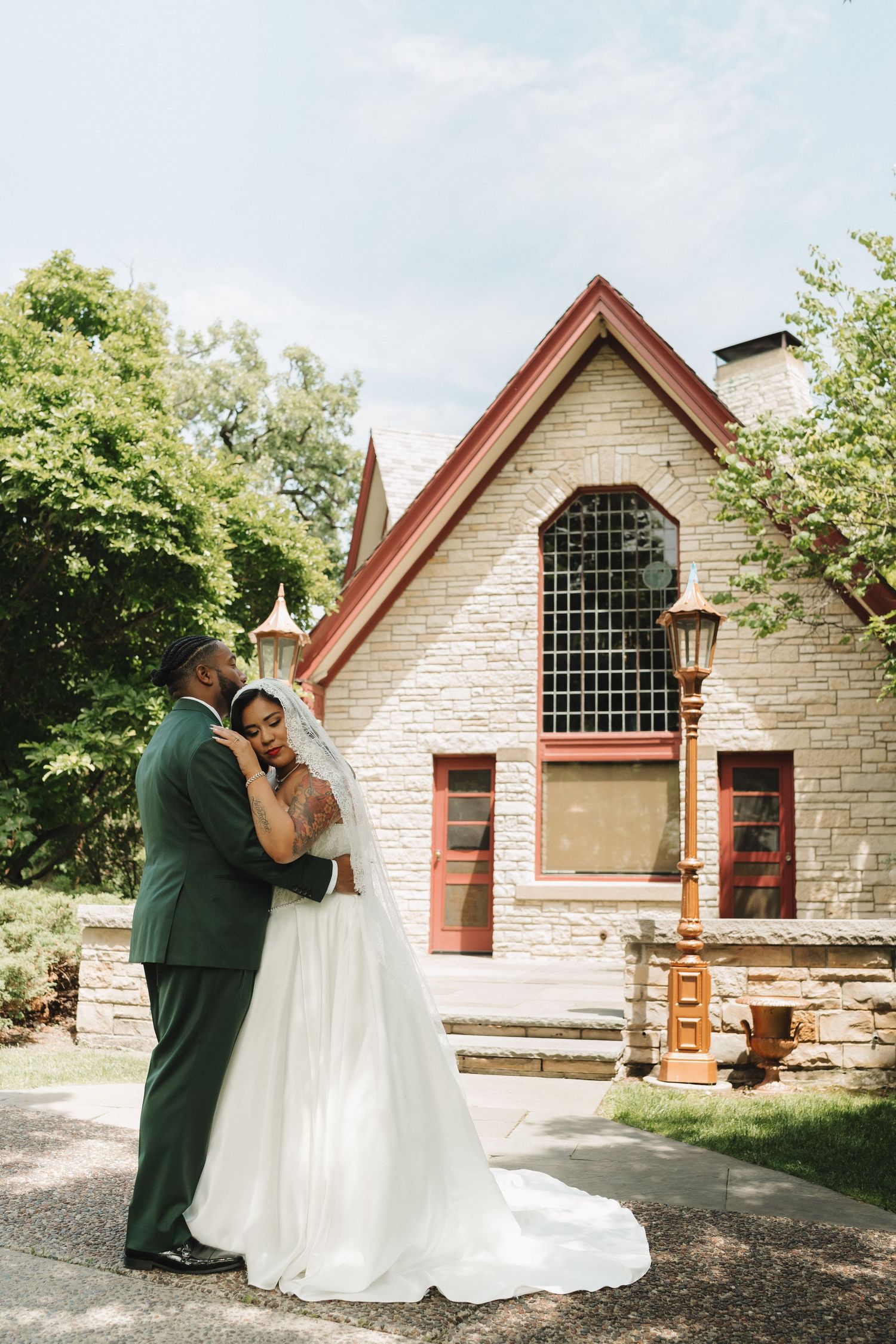 A bride and groom embrace outside a charming stone chapel with red trim on their wedding day.