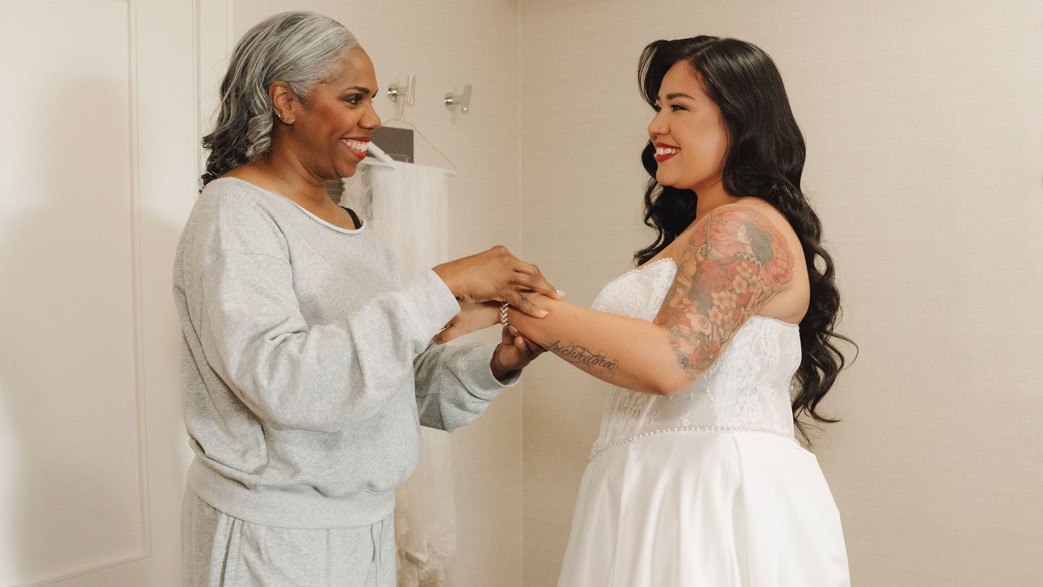 Bride in white wedding dress holds hands with older woman in blue outfit in bright room.
