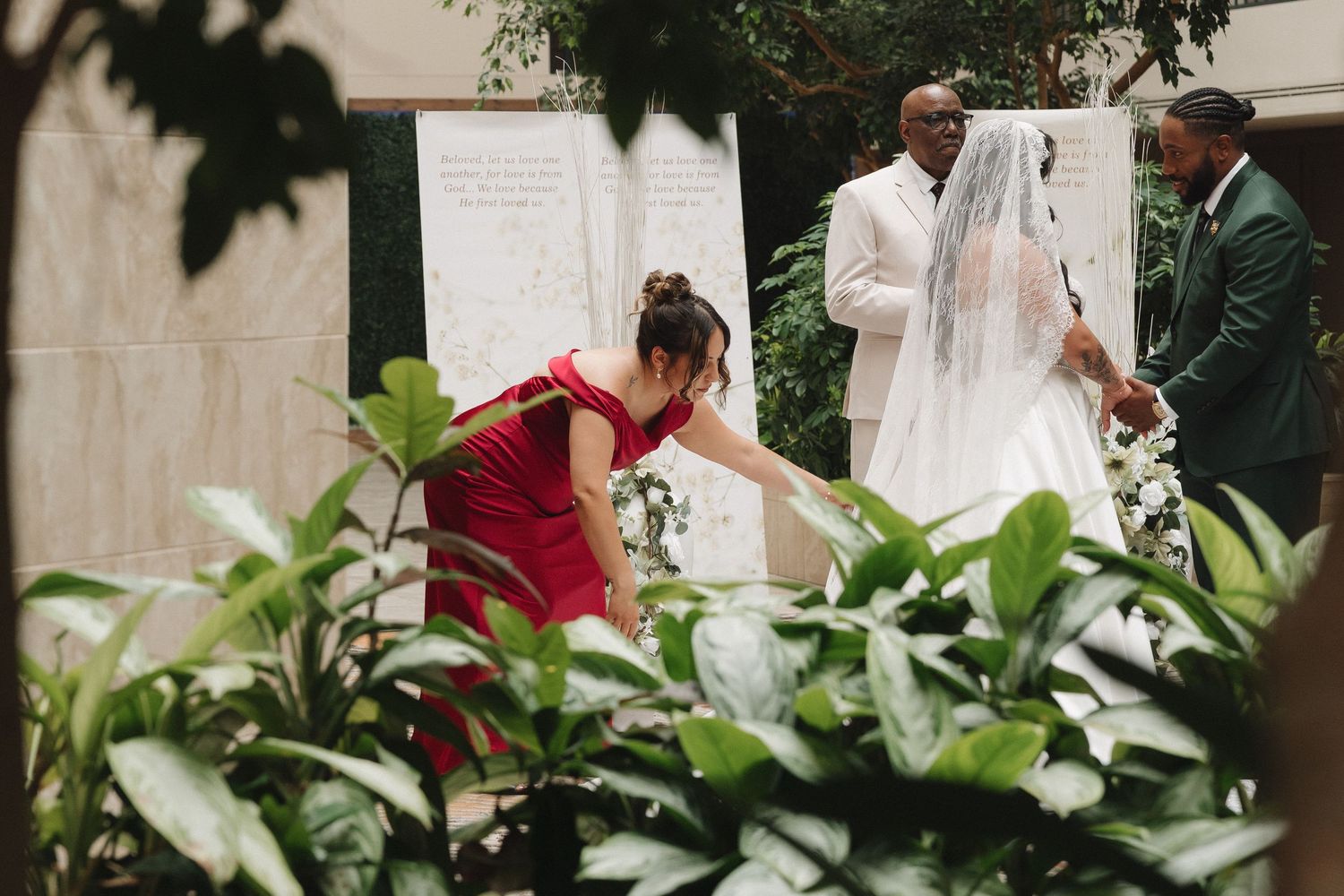 A bride in a white gown and veil stands with guests while a woman in red adjusts her dress at an elegant indoor wedding.