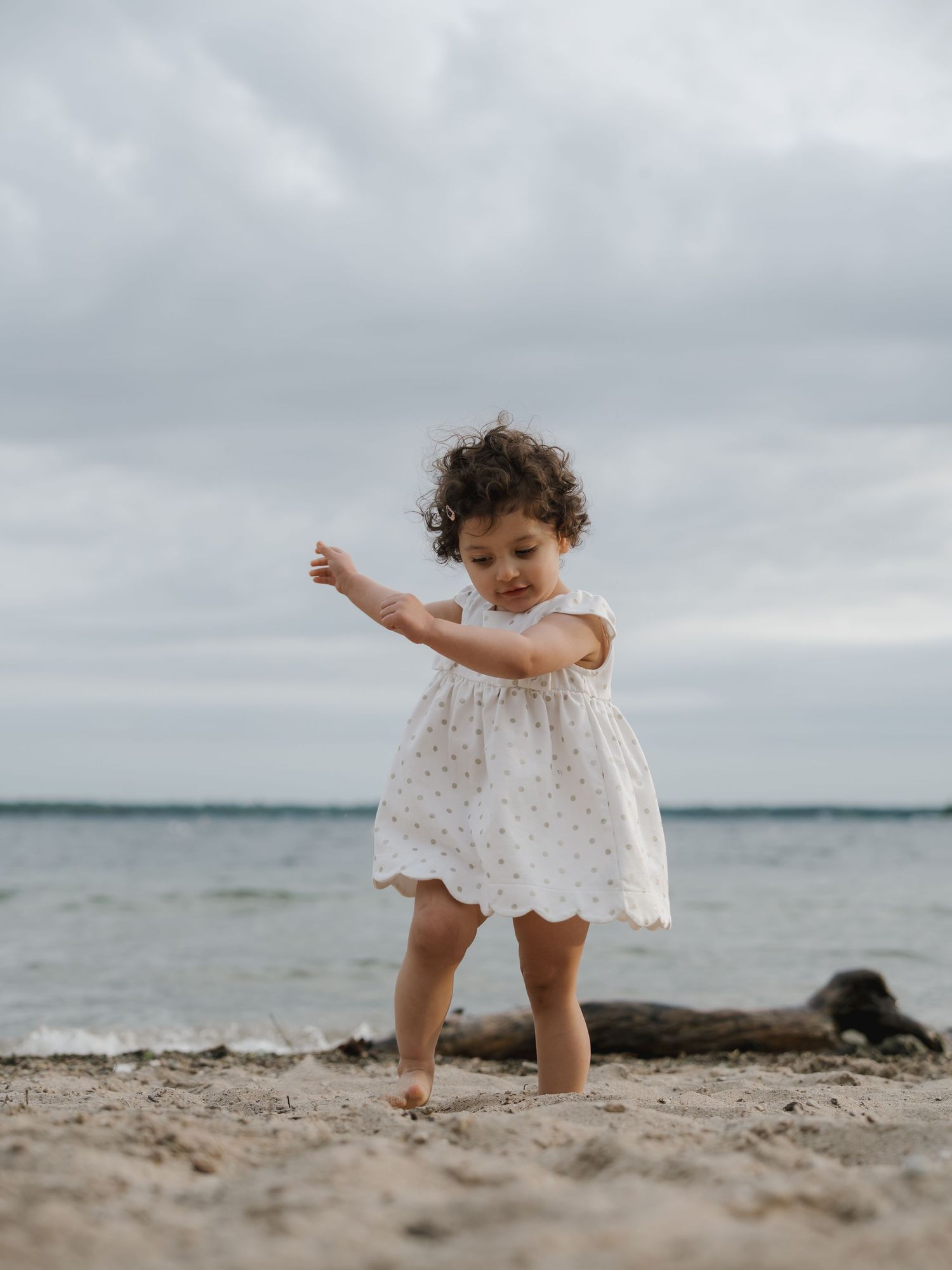 A toddler girl in a white floral dress walks barefoot on a sandy beach under a cloudy sky.