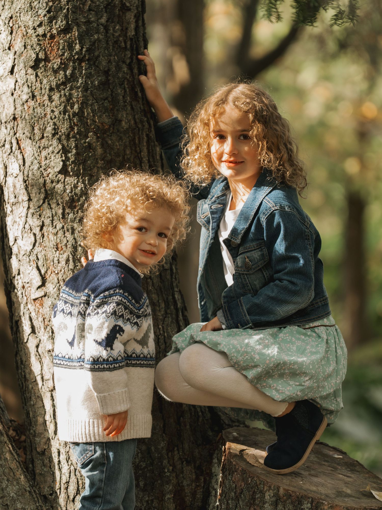 Two curly-haired children pose by a tree in a forest, wearing cozy sweater and denim jacket.