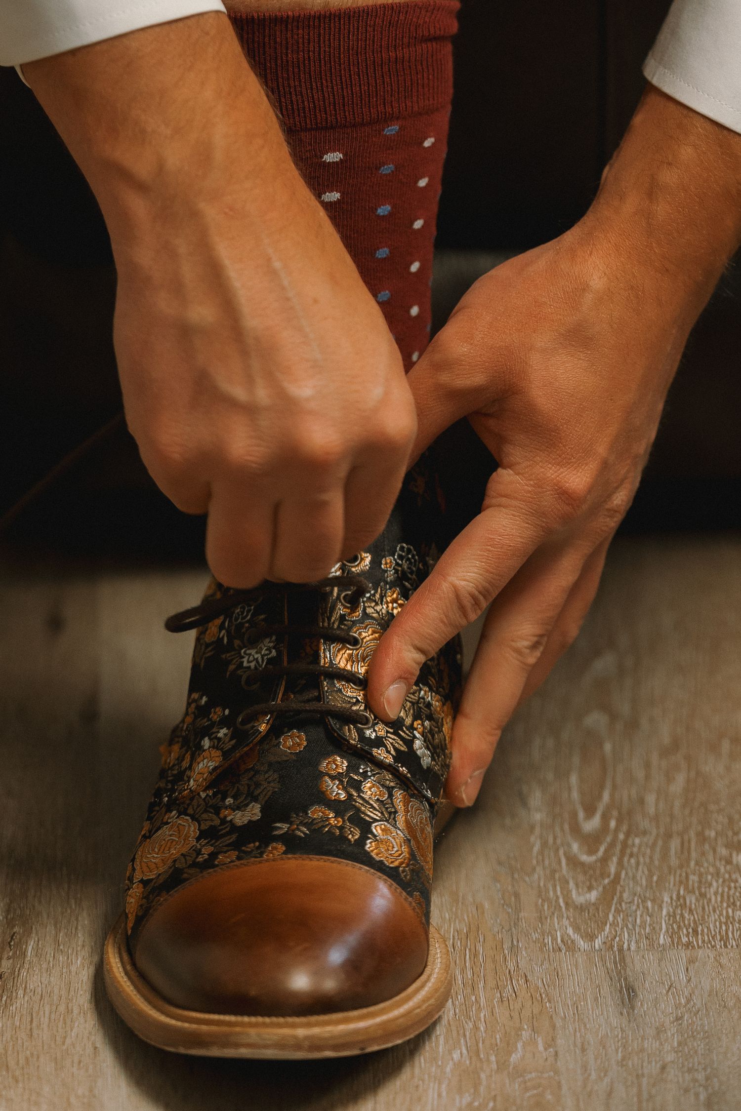 Groom tying laces on decorative floral brocade dress shoes with colorful polka dot socks.