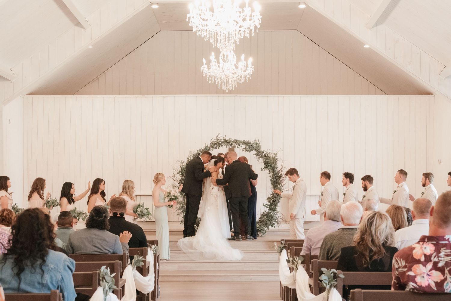 Bride and groom exchange vows at floral arch altar in white chapel with wedding party.