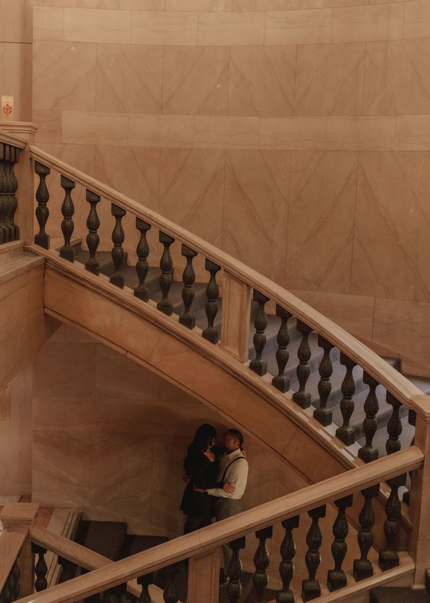 Bride and groom embracing on elegant curved marble staircase with ornate balusters.