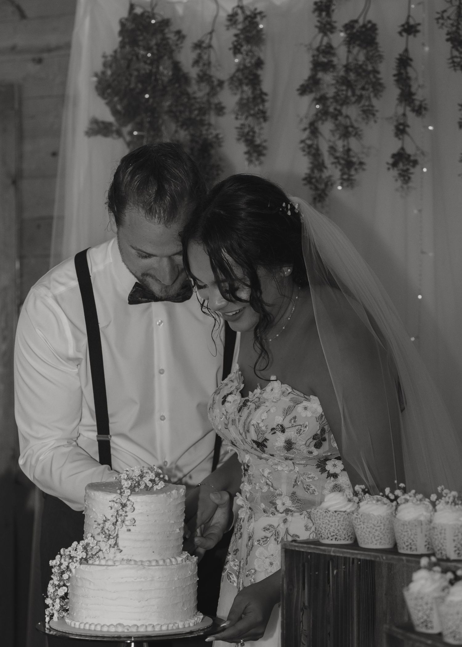 Black and white photo of smiling bride and groom cutting their two-tier wedding cake at their reception.