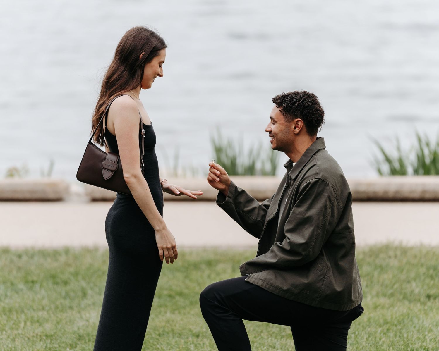 Man proposing on one knee to woman in black dress outdoors near water on green grass.