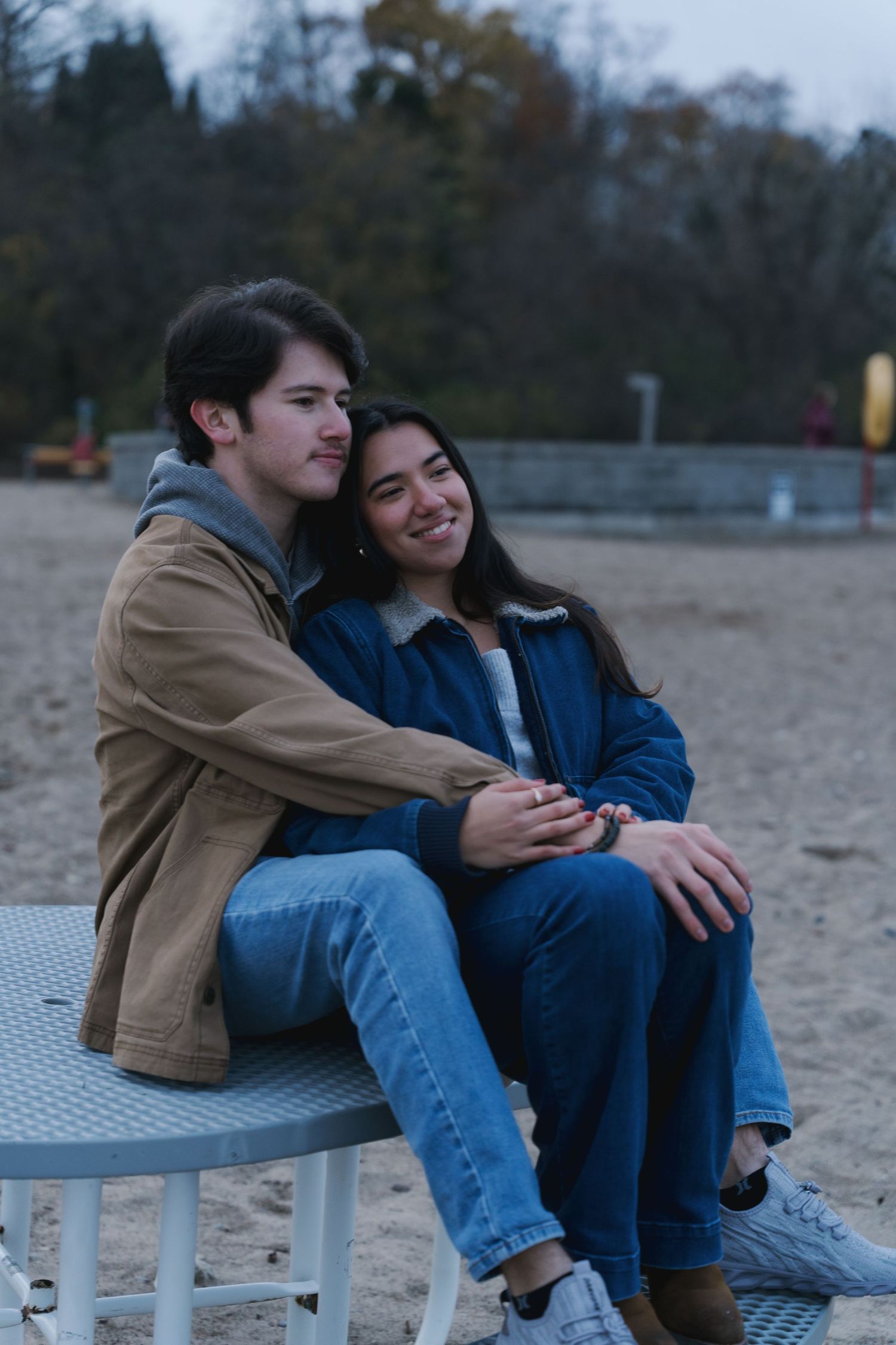 A happy couple sits closely together on a table at a sandy beach in autumn.