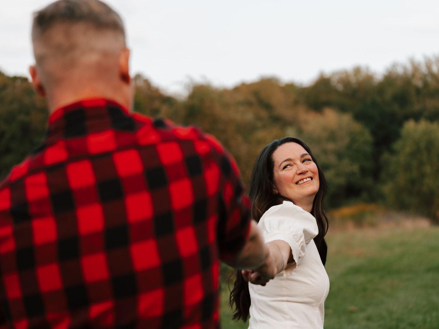 Smiling dark-haired woman holds her partners hand while walking through a green meadow.