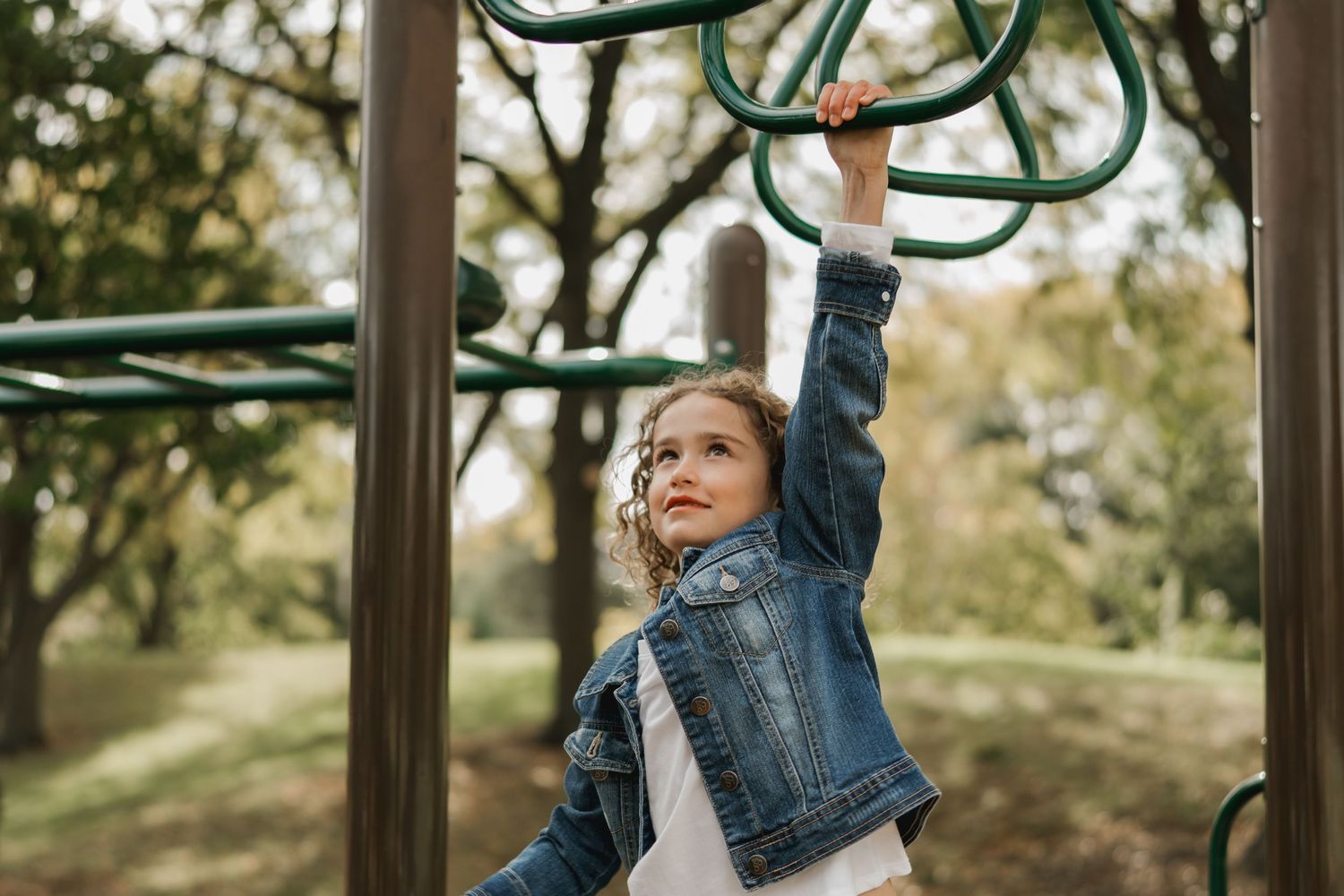 Young girl in denim jacket hangs from monkey bars at an outdoor playground.