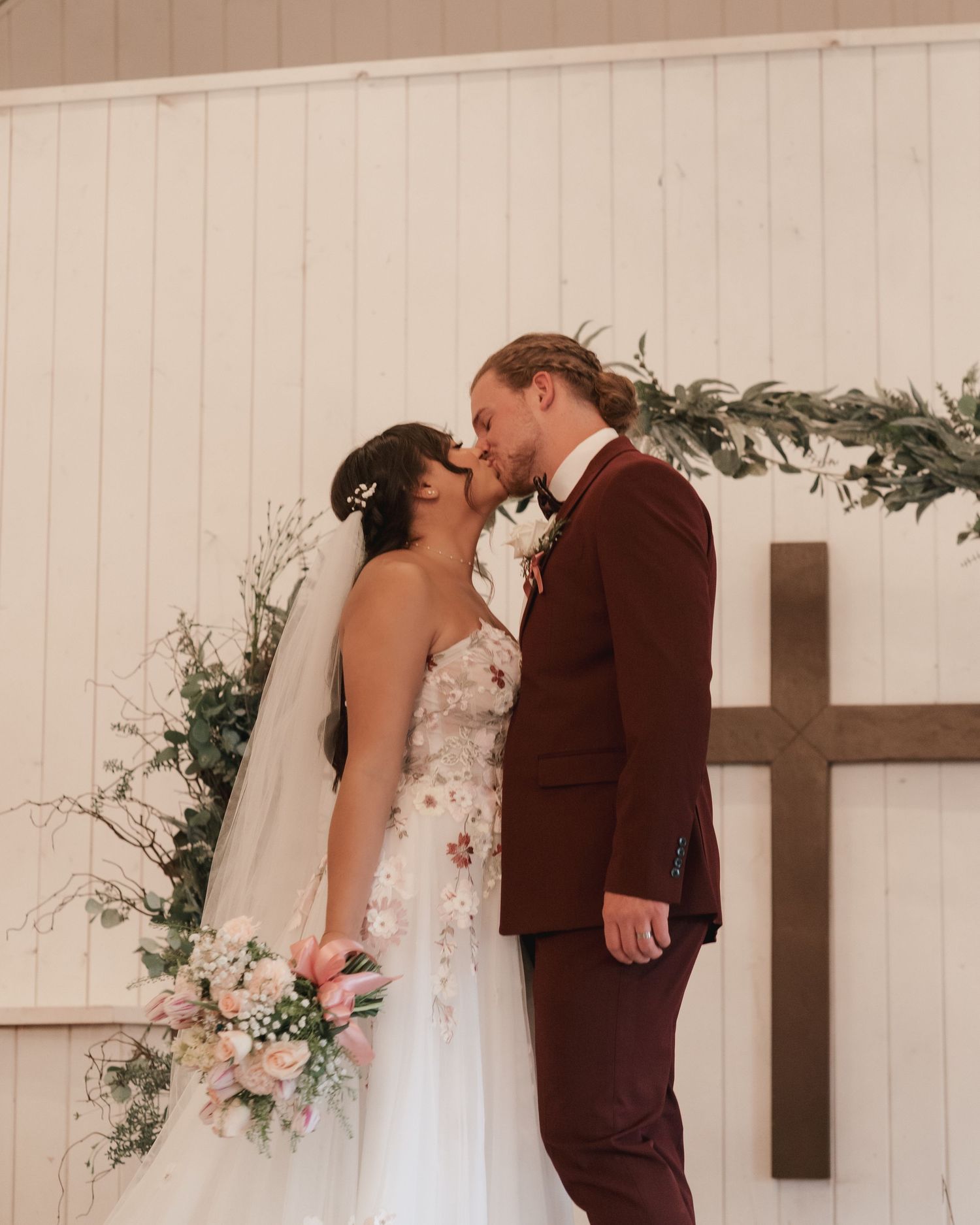 Newlyweds kiss in front of a wooden cross and floral arch in a white chapel wedding venue.