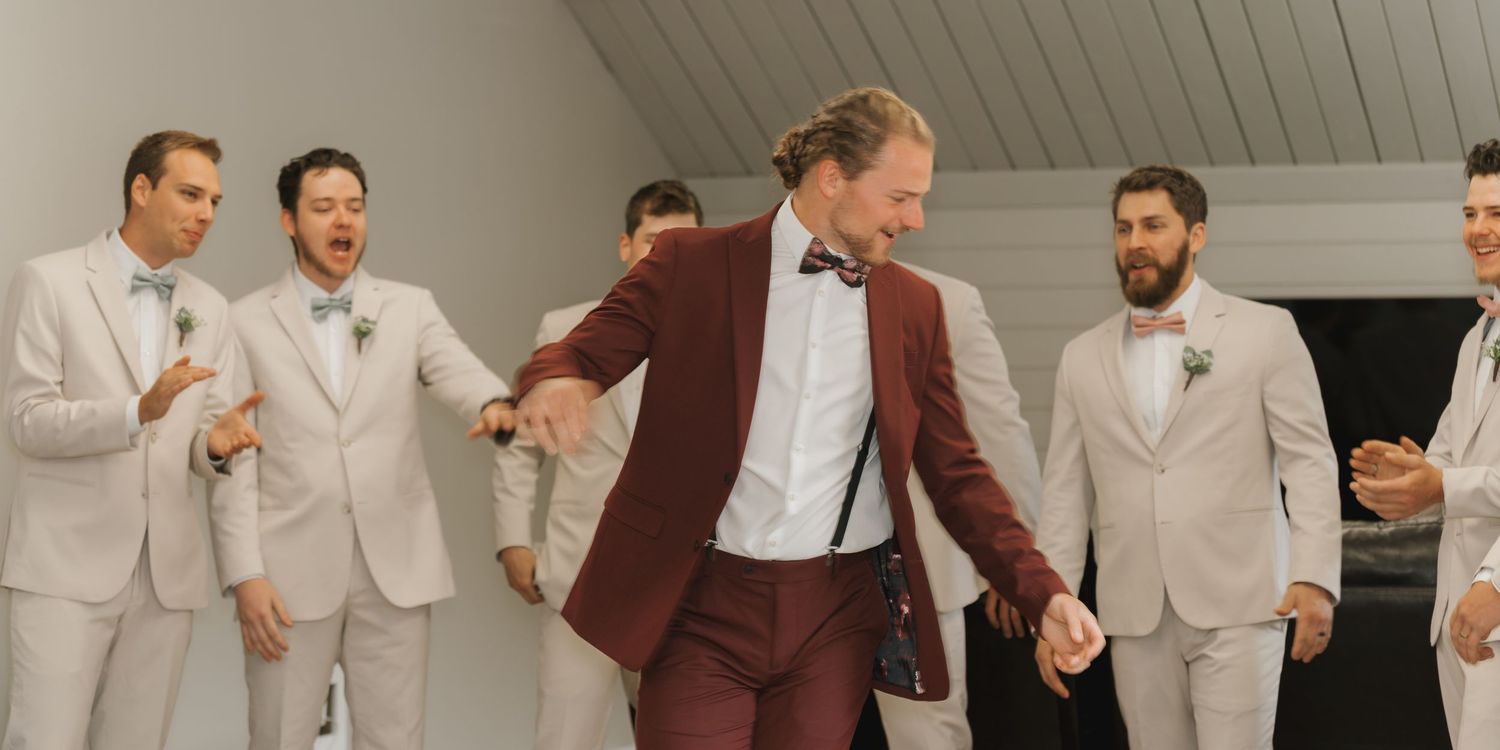Groom in burgundy suit dancing joyfully with groomsmen in light gray suits at wedding reception.