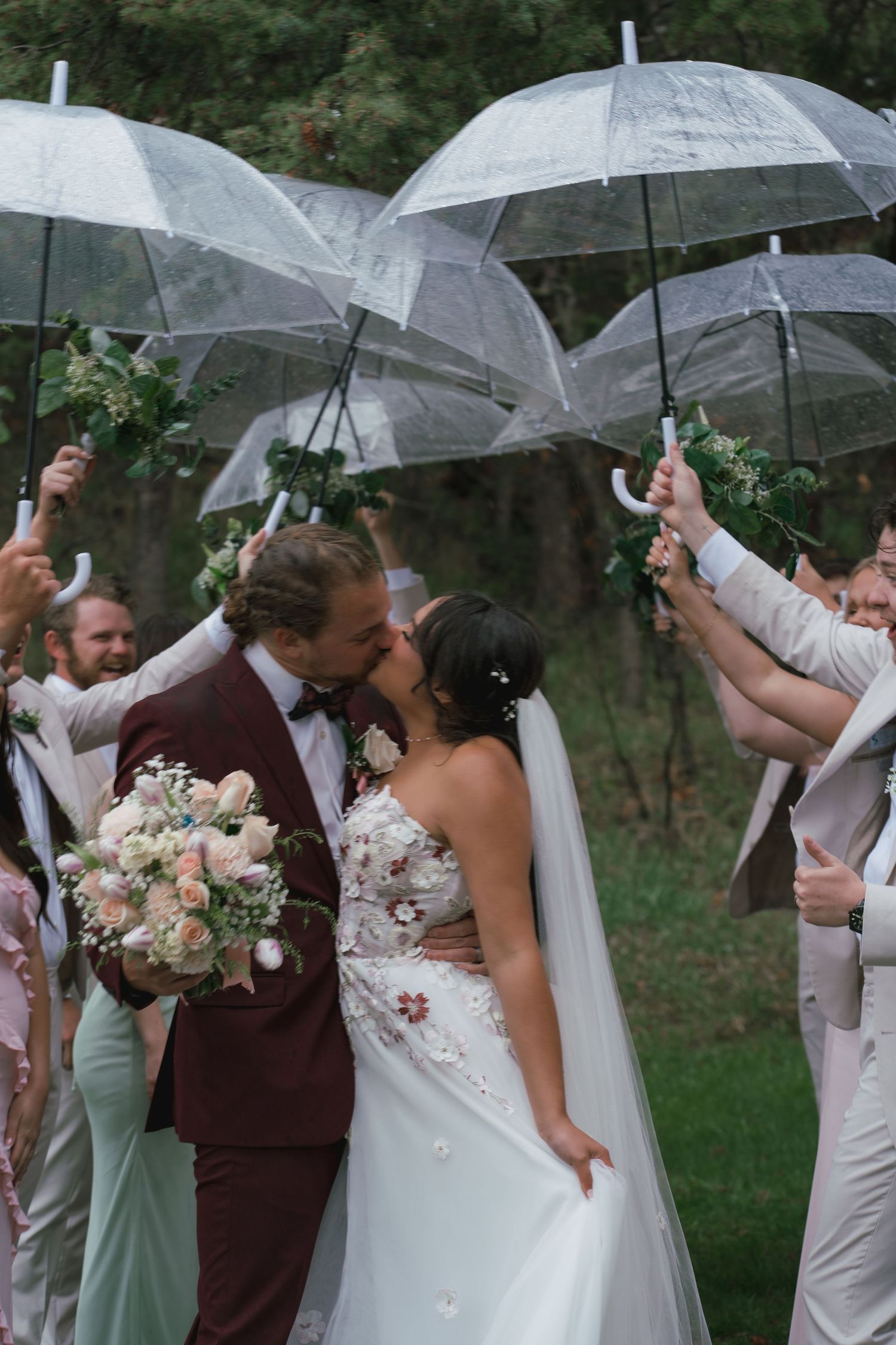 Bride and groom share a kiss under clear umbrellas held by wedding party outdoors, bride holding blush bouquet.