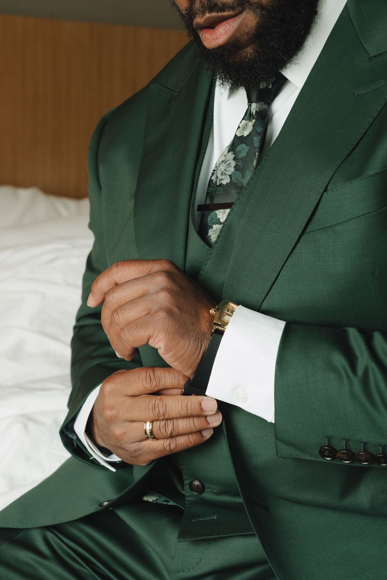 Man in green three-piece suit adjusting cufflinks, wearing gold watch and ring, with floral tie.