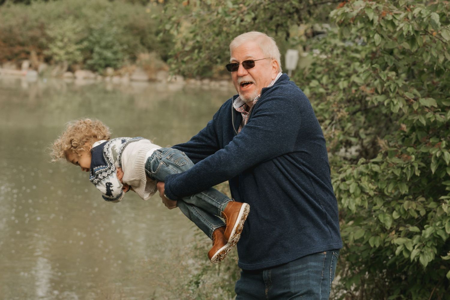 A grandfather in navy jacket playfully lifts a small child near a pond outdoors.