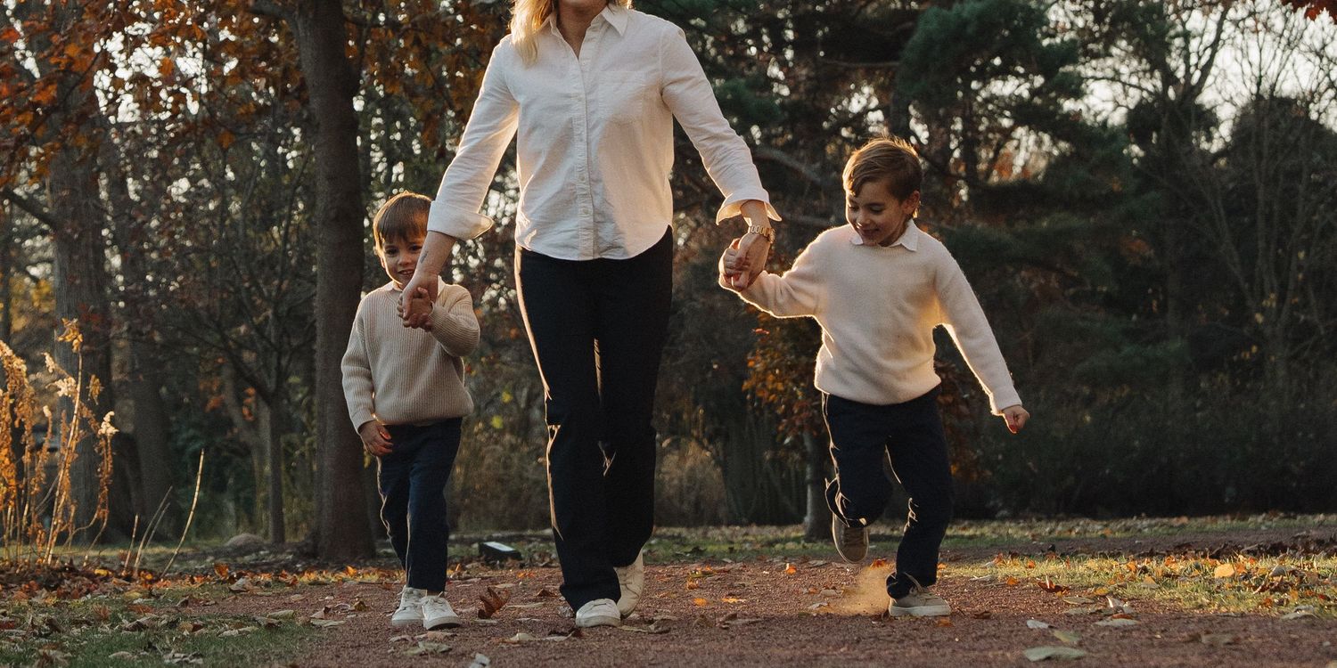 Mother walks hand-in-hand with two young children along a sunlit autumn park path.