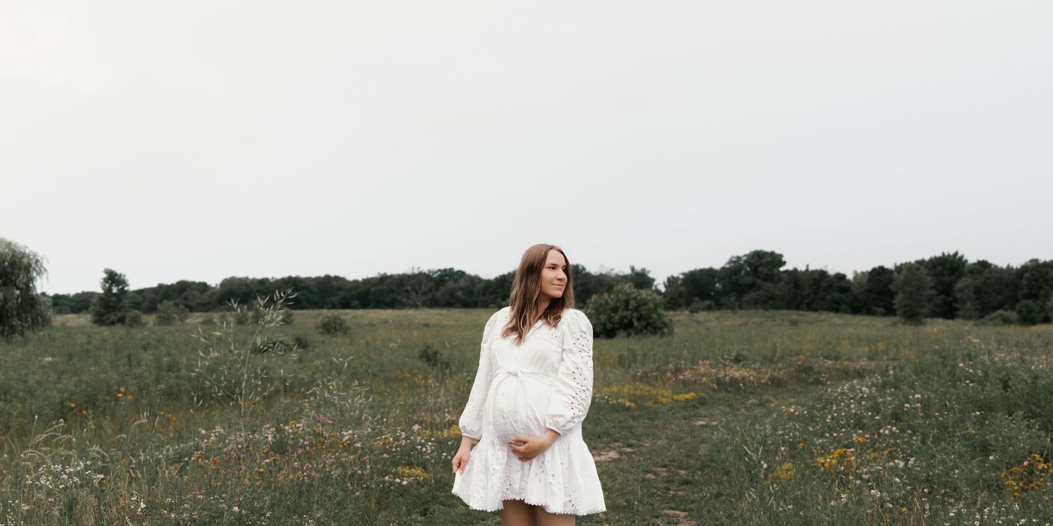 Pregnant mother in white dress standing in a wildflower meadow on an overcast day.