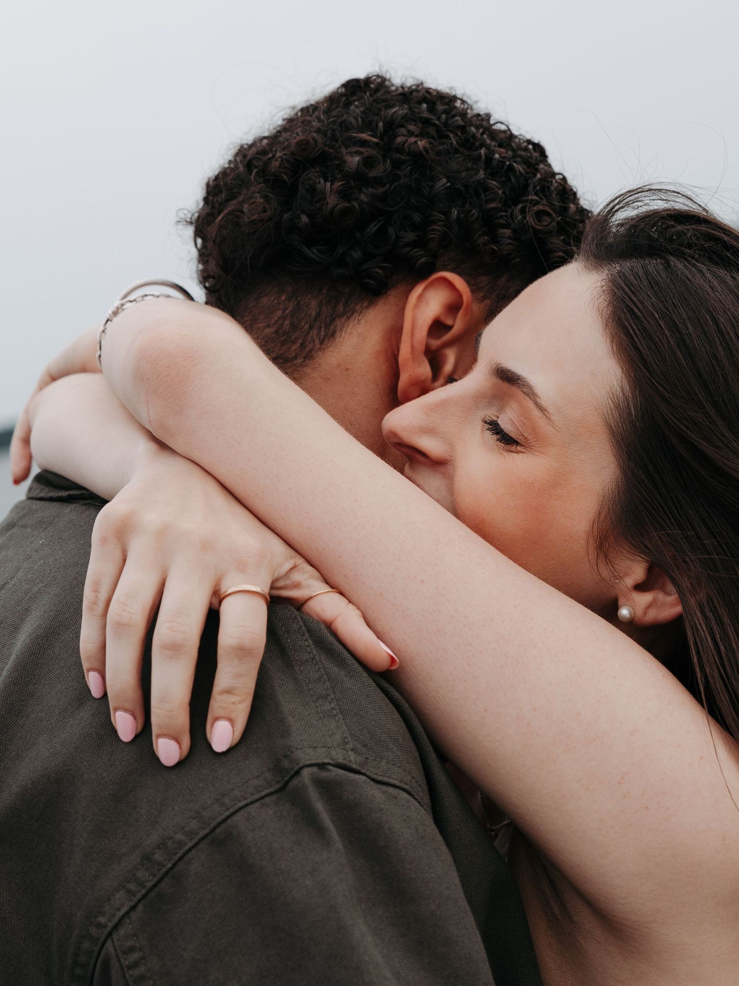 A couple sharing a tender embrace, woman with pink nails and ring hugging man in olive shirt against soft sky background.