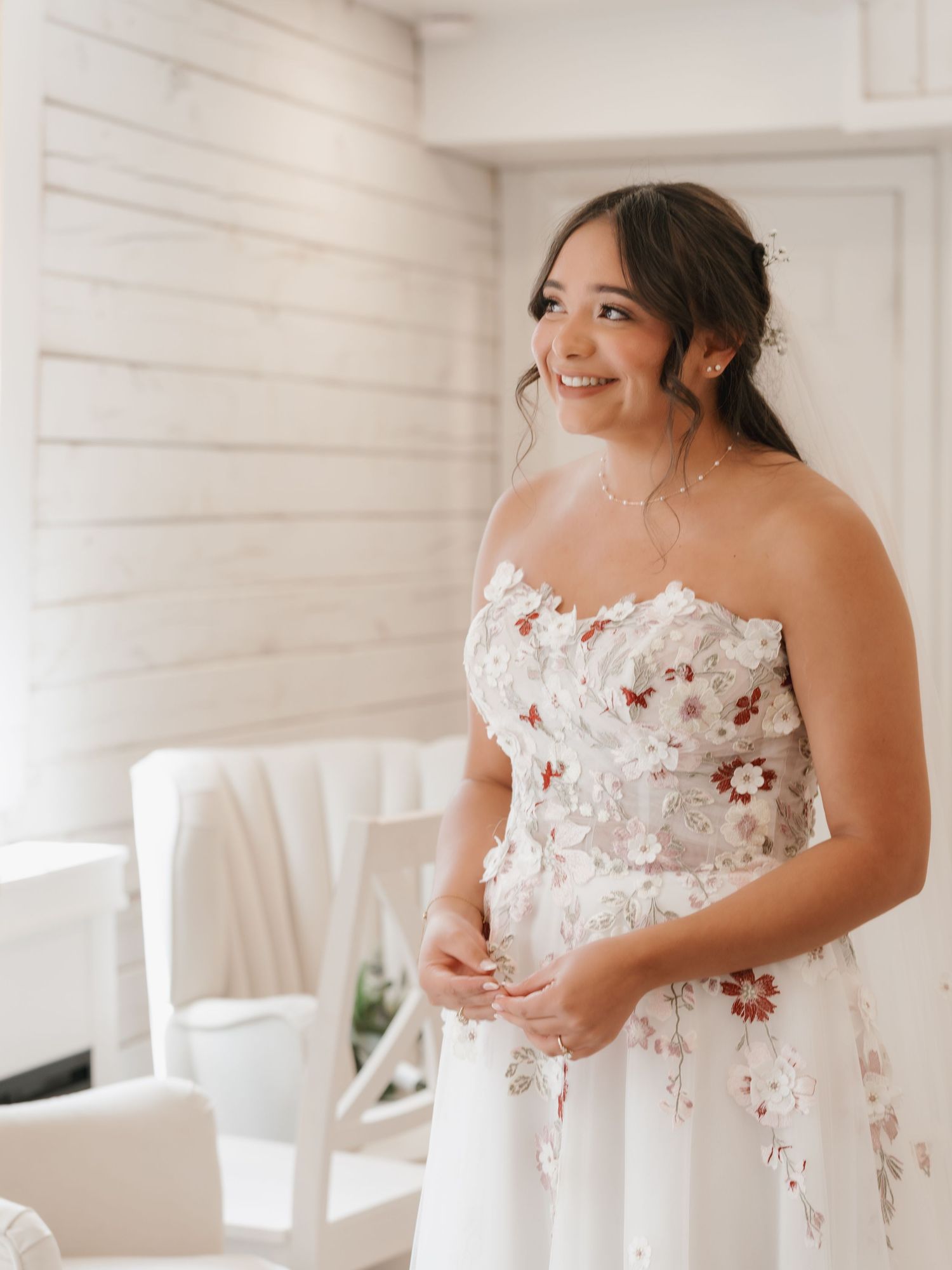 Smiling bride in a strapless white gown with floral appliques stands in a bright white room on her wedding day.
