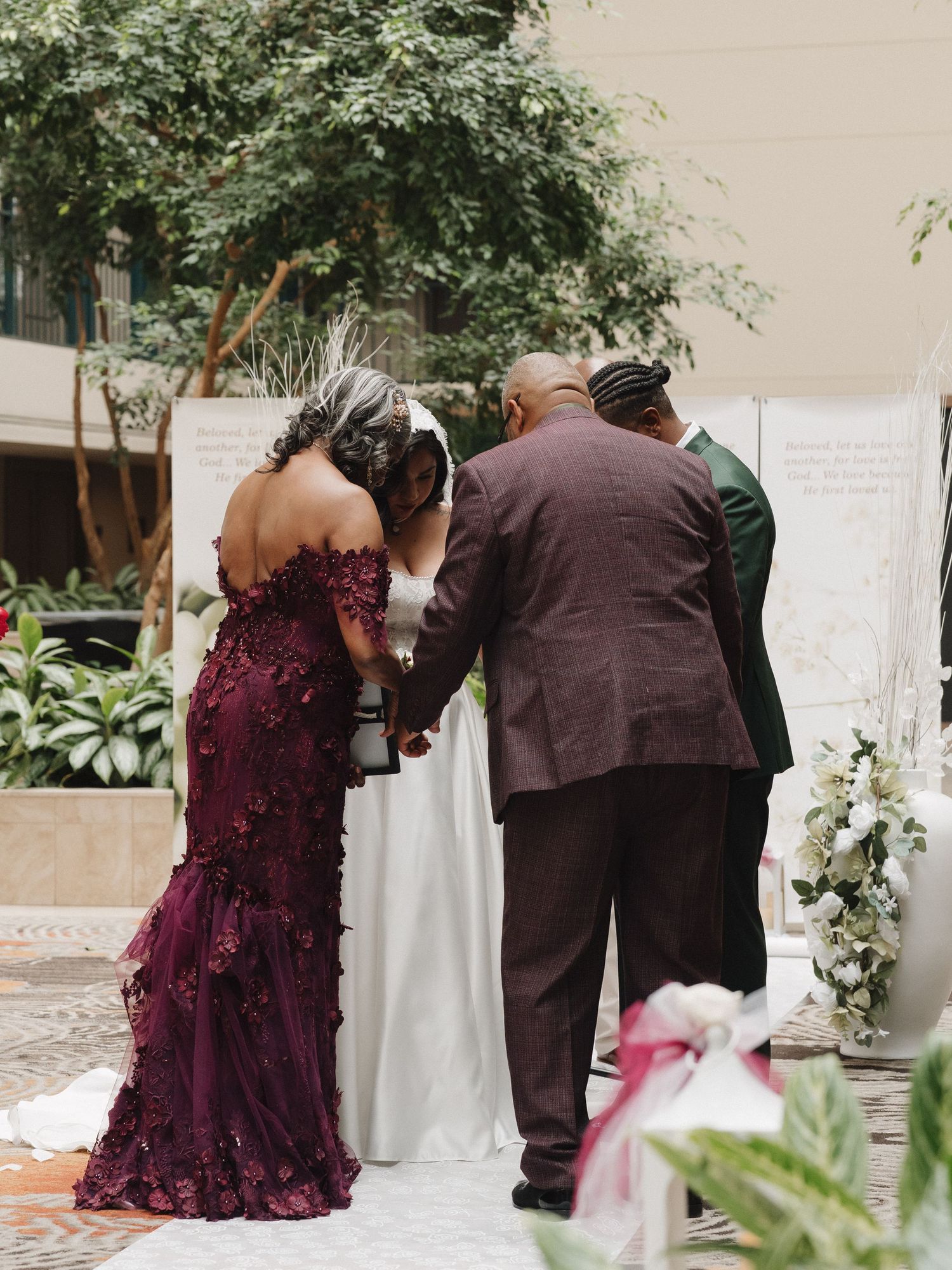 Wedding ceremony with bride in white gown, groom in burgundy suit, and guest in floral dress holding hands indoors.