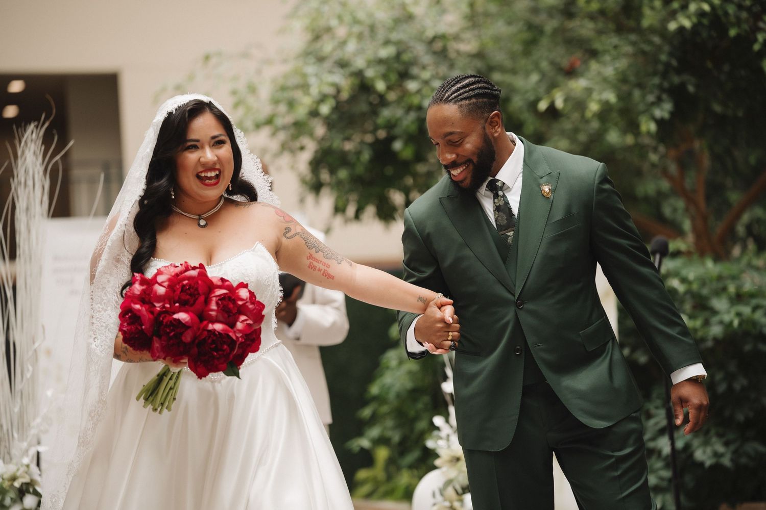 Joyful bride in white gown with red bouquet and groom in green suit celebrating after wedding ceremony.