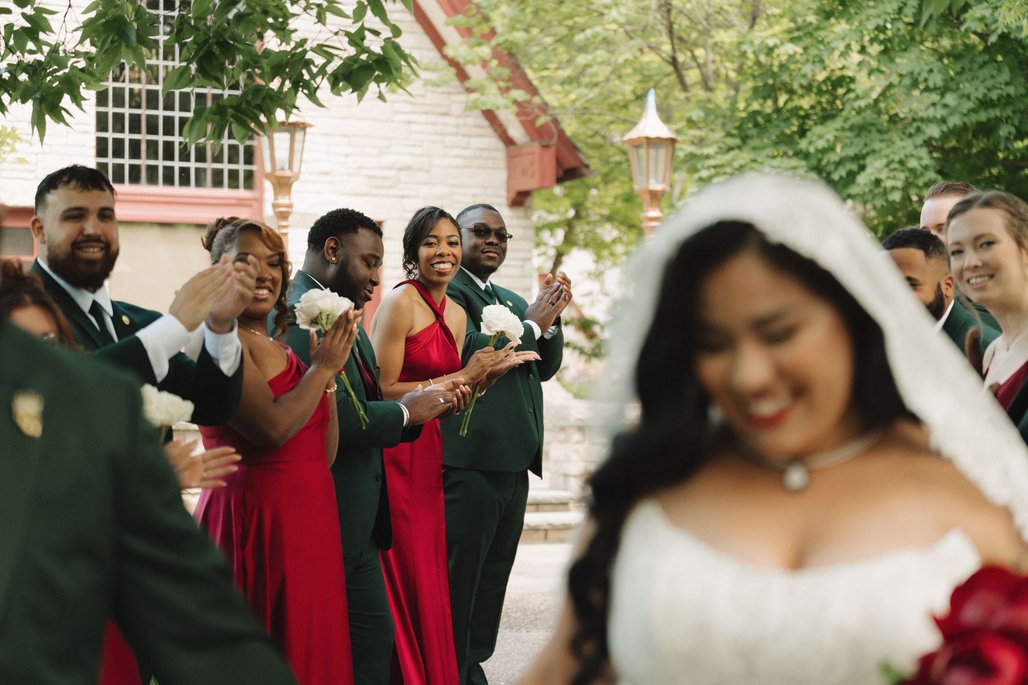 Wedding party in red and green attire cheers as bride in white gown and veil smiles in foreground.