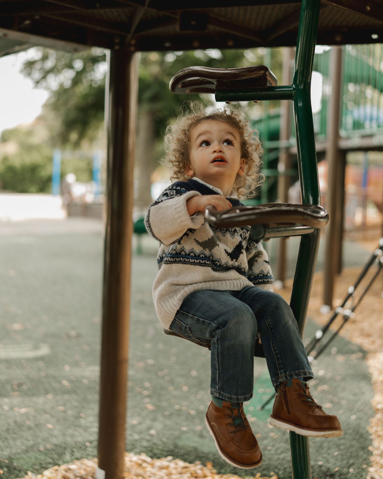 Curly-haired toddler in a patterned sweater plays on playground equipment outdoors.
