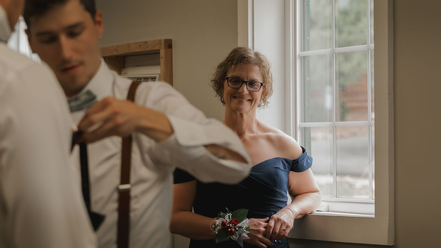 Mother of the groom smiling warmly while watching groom adjust tie near bright window.