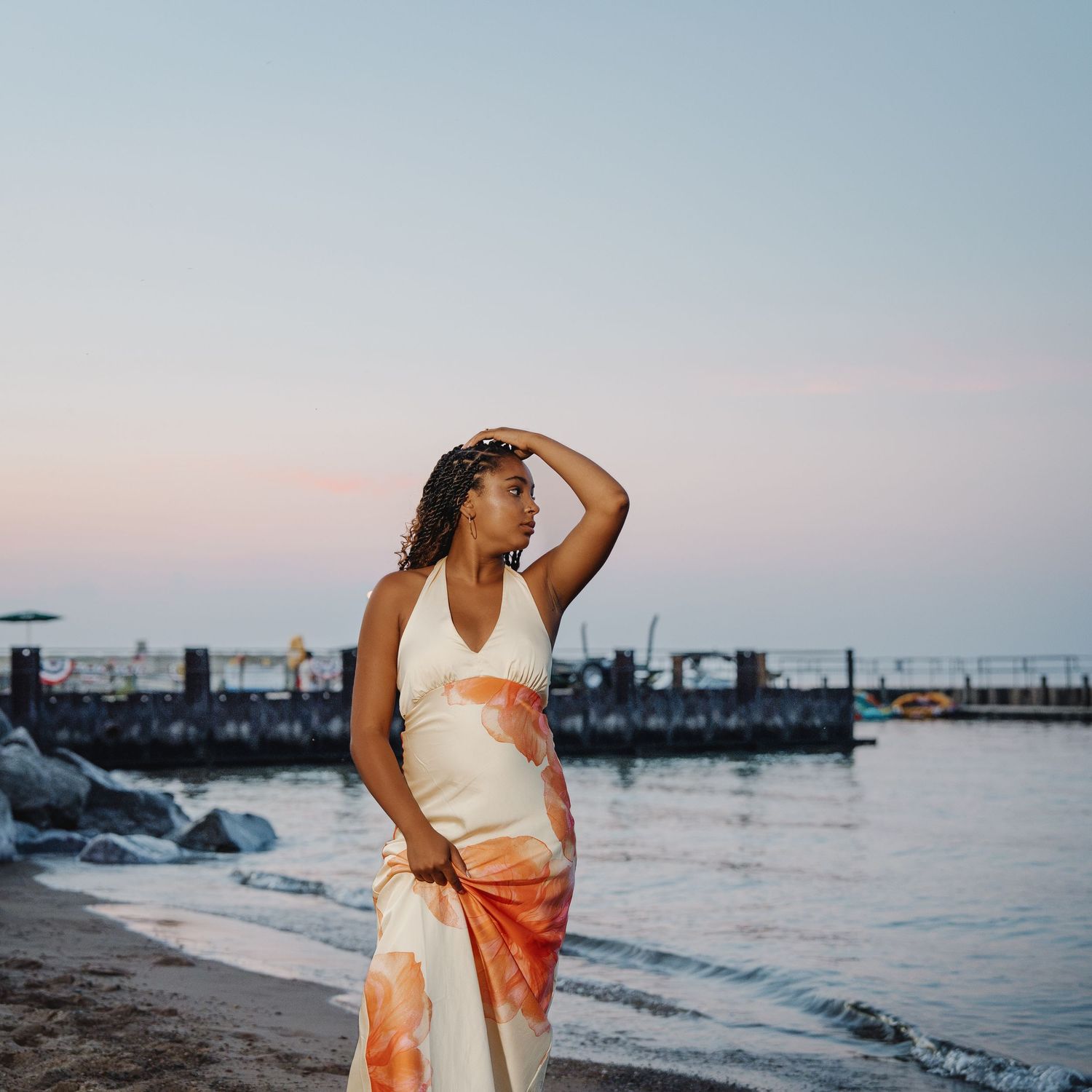 Woman in white and orange tie-dye dress poses on sandy beach at sunset with calm water and pier in background.