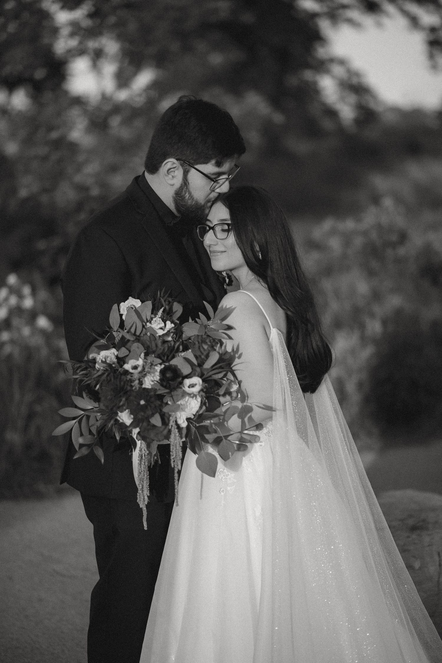 Black and white wedding photo of bride holding bouquet and groom tenderly kissing her forehead outdoors.