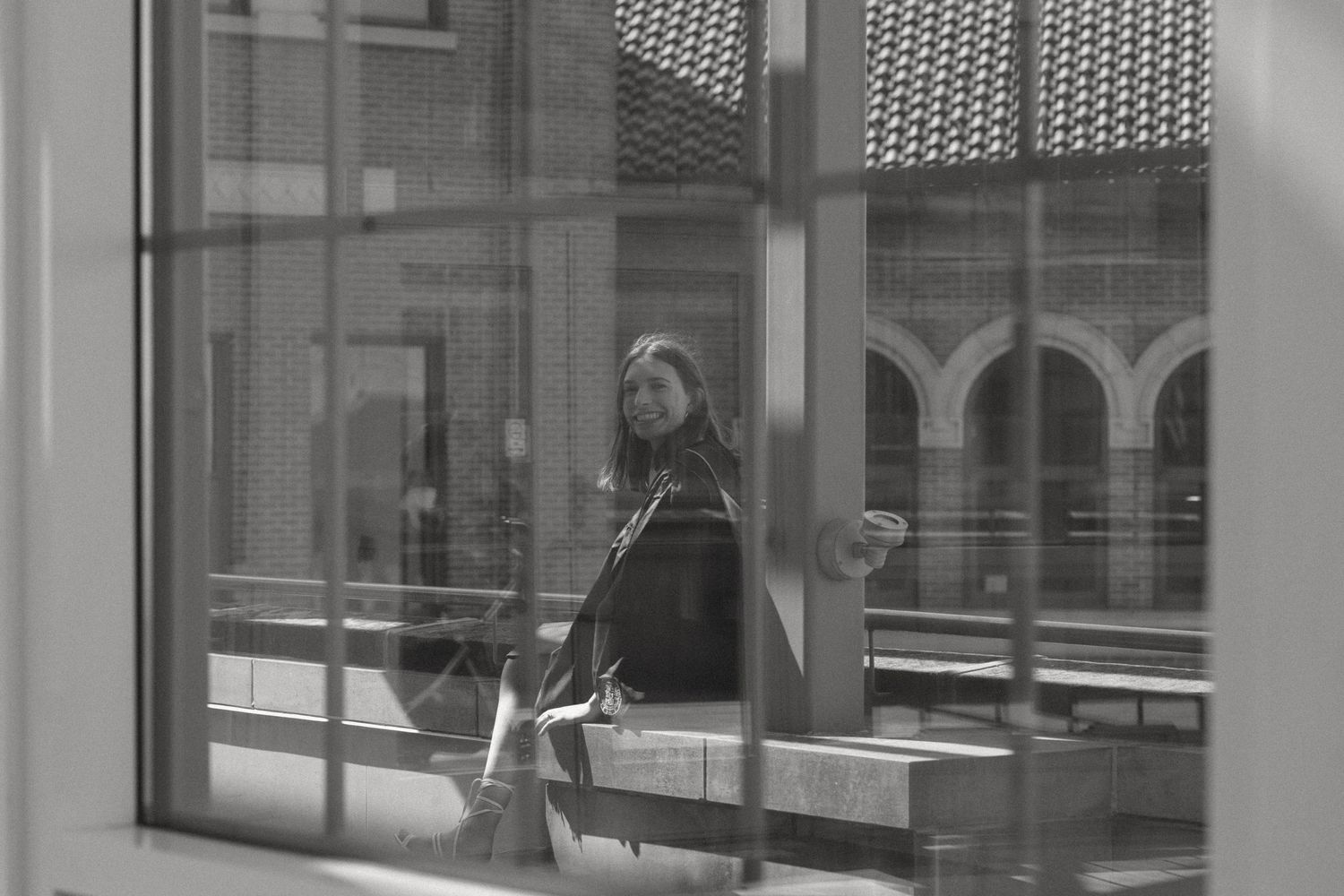 Black and white photo of a woman sitting on a bench, viewed through a large window of a modern building.
