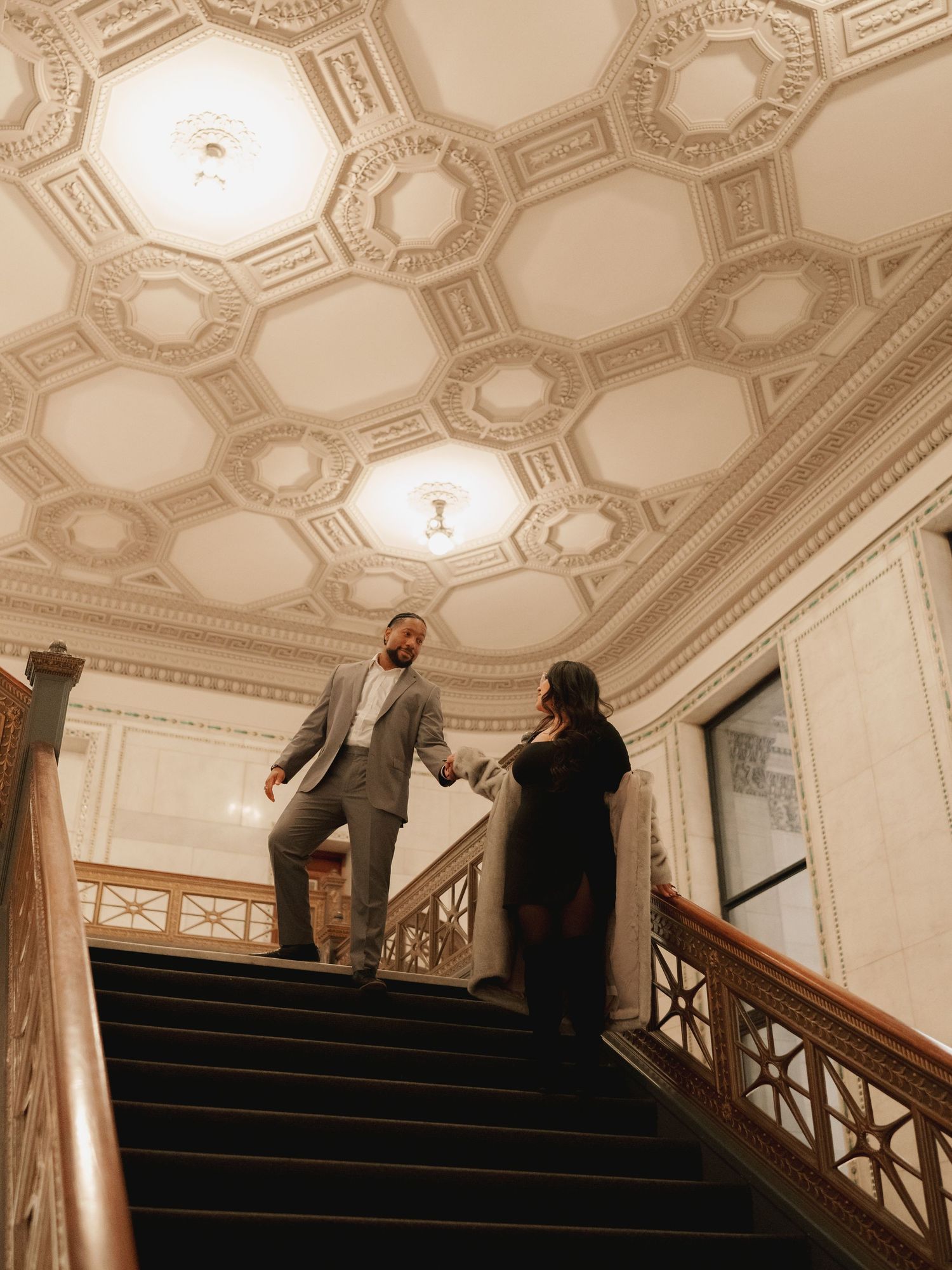 Couple holds hands descending grand staircase beneath ornate coffered ceiling in historic building.