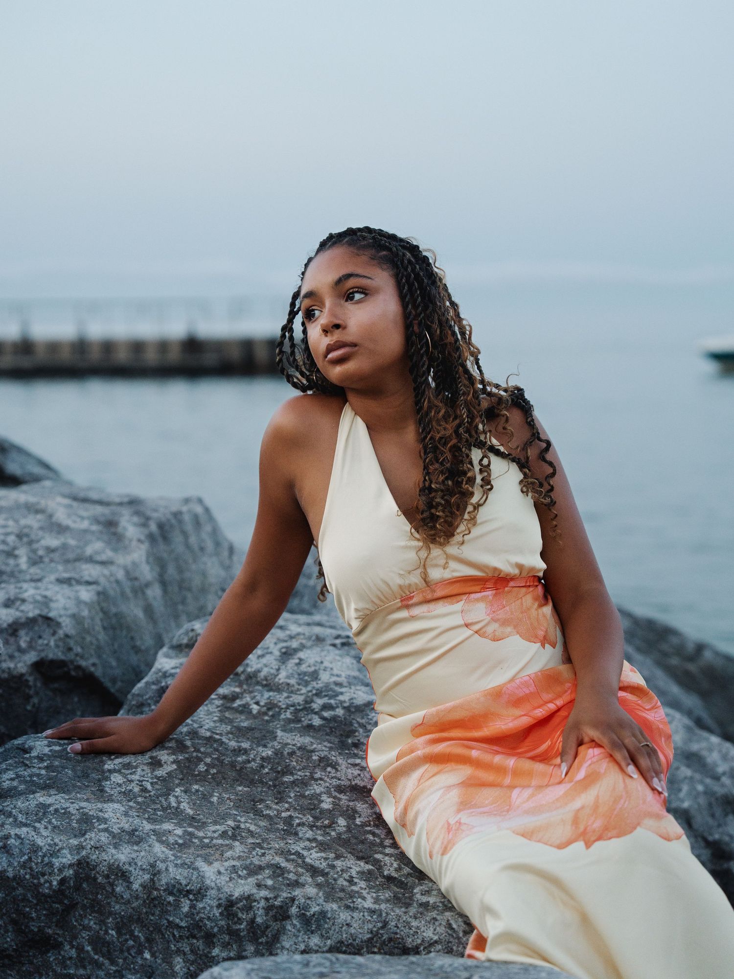 Young woman in colorful halter maxi dress sitting on rocks by the ocean at dusk.