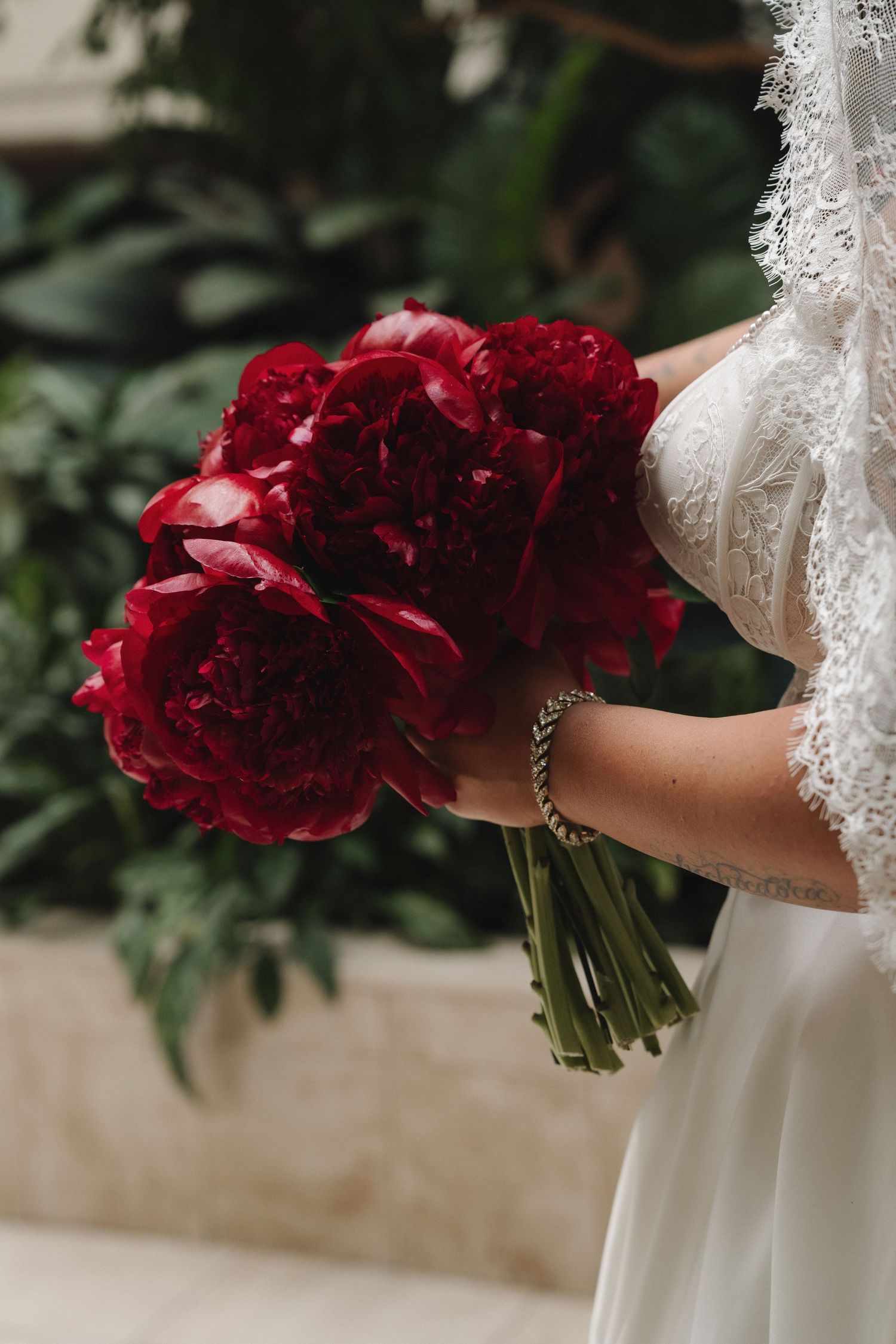 Bride in white lace gown holding a lush bouquet of deep red peonies with green stems at a wedding ceremony.
