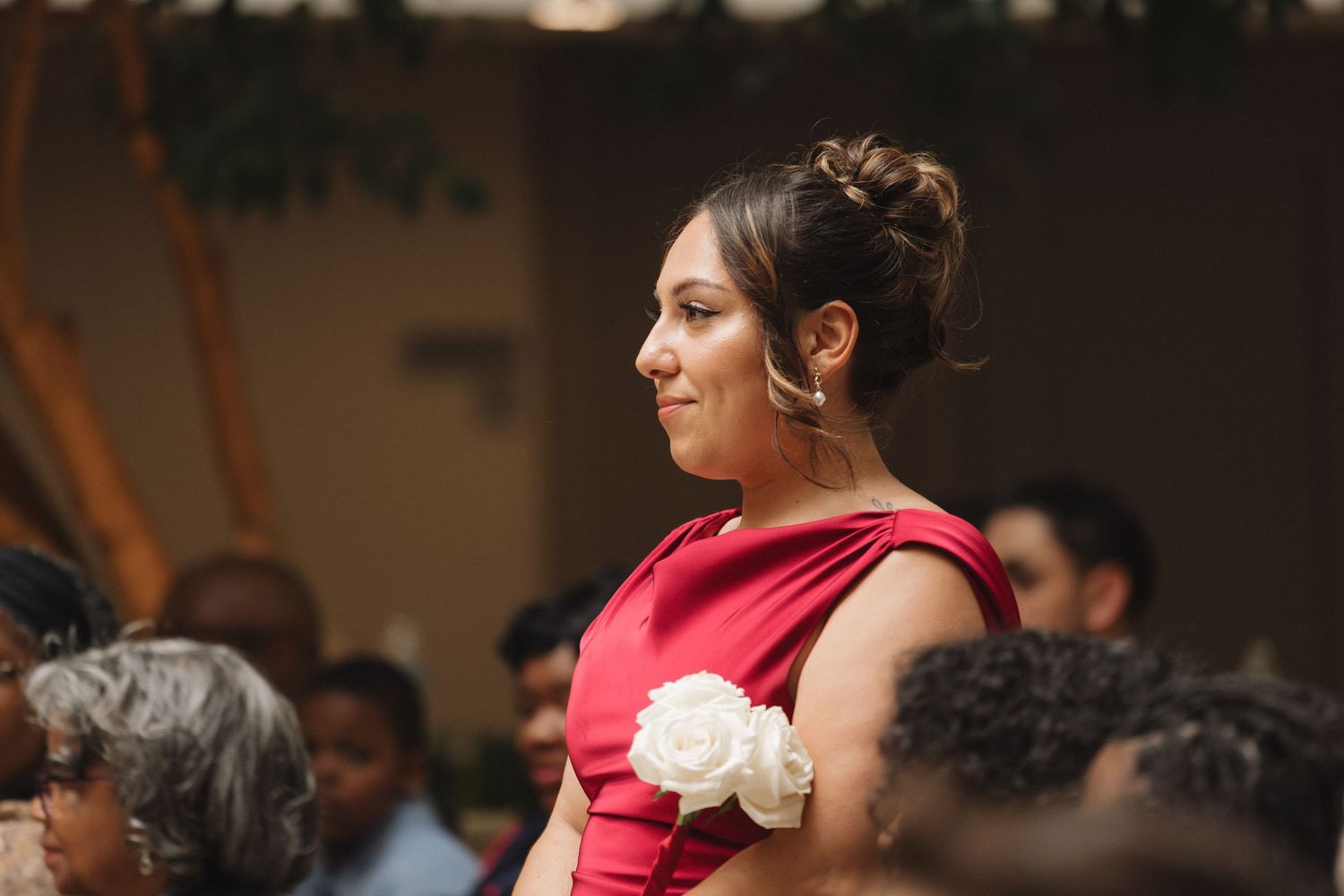 Woman in red dress with white rose corsage stands elegantly at a formal event, smiling softly in a warmly lit venue.