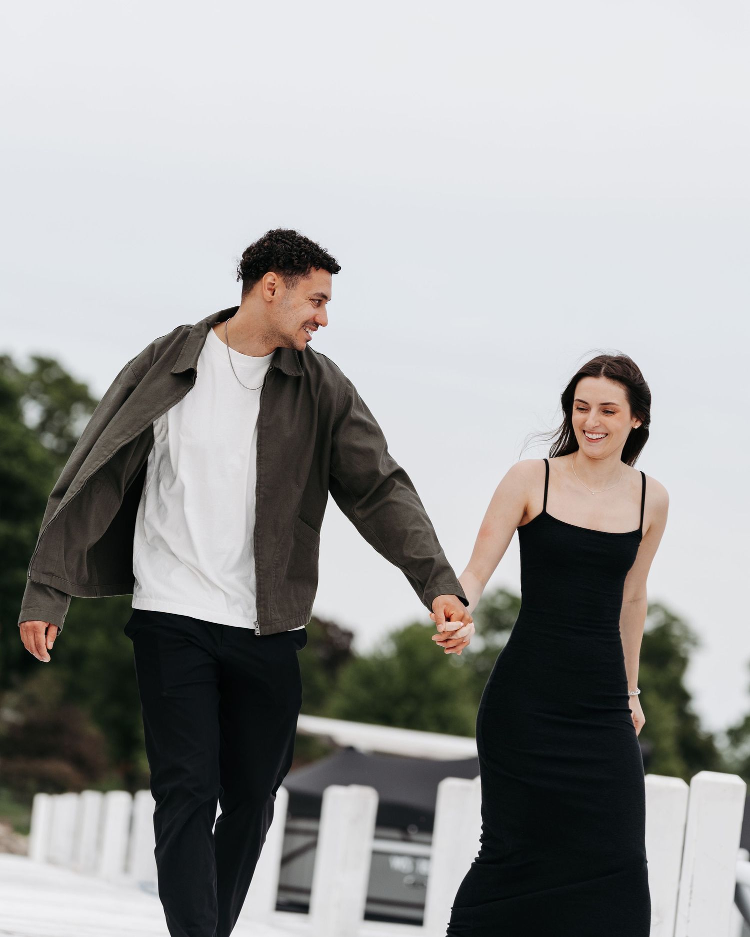 A smiling couple walks hand in hand outdoors, man in olive jacket and white tee, woman in elegant black dress.
