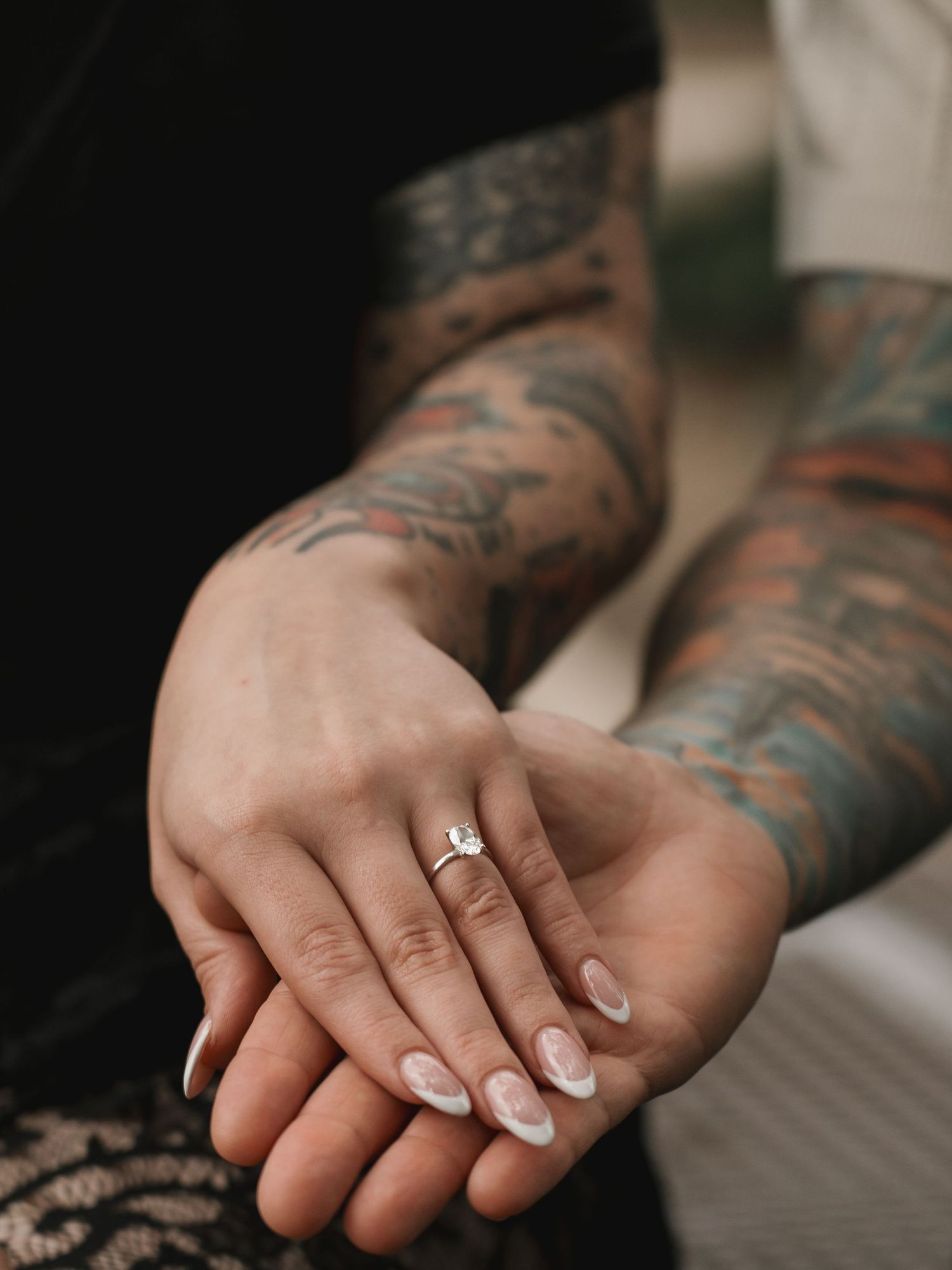 Close-up of hands with French tip nails and an engagement ring resting on heavily tattooed arms.