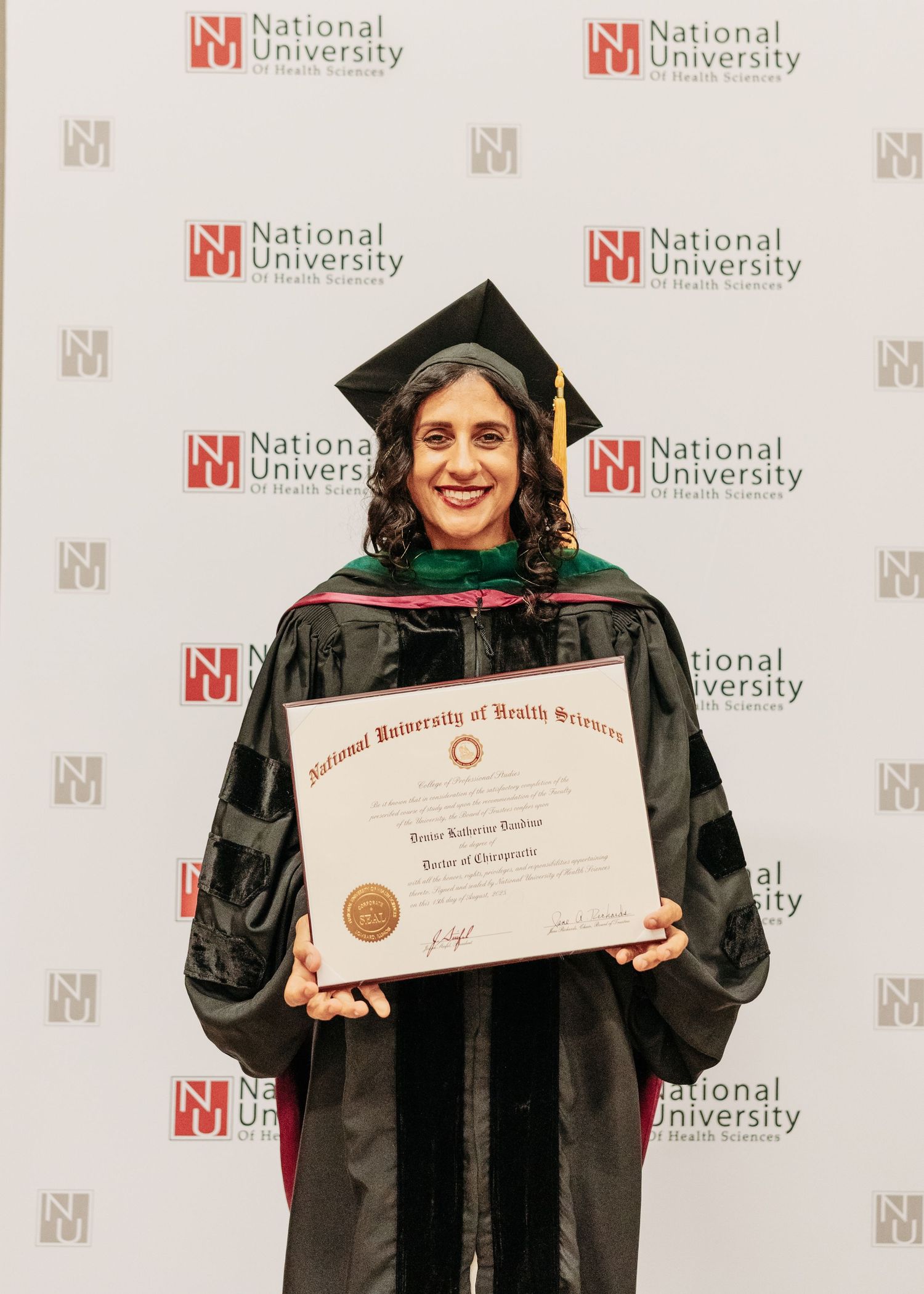 Graduate in black cap and gown holds diploma at National University of Health Sciences commencement ceremony.