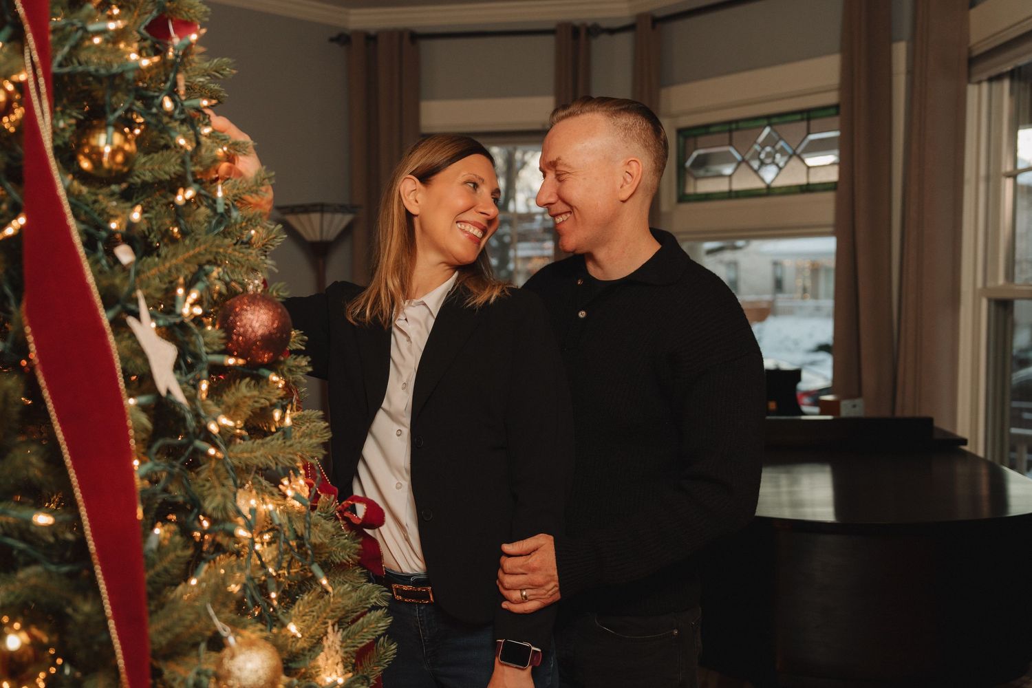 Couple smiling at each other beside a decorated Christmas tree with red ribbon and ornaments indoors.